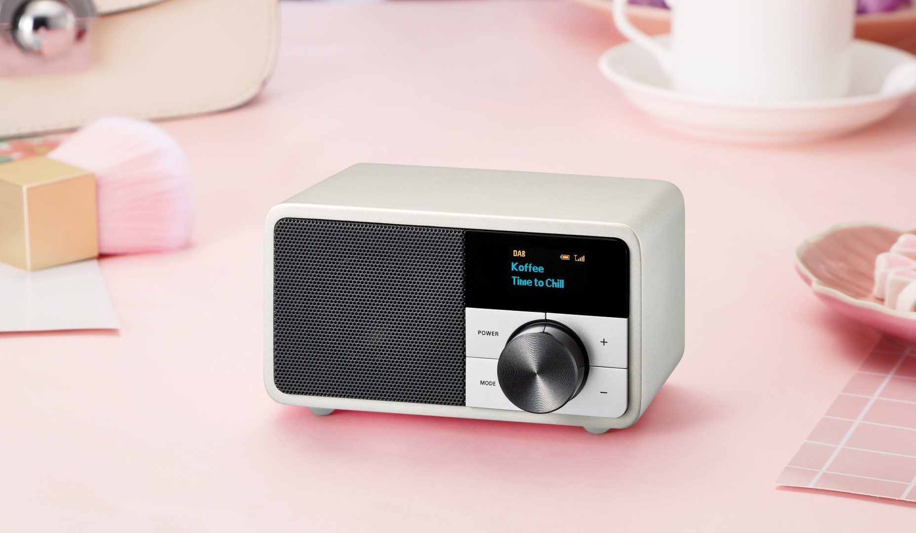 Radio blanche avec un écran affichant 'Koffee' et 'Time to Chill'. Posée sur une surface rose.