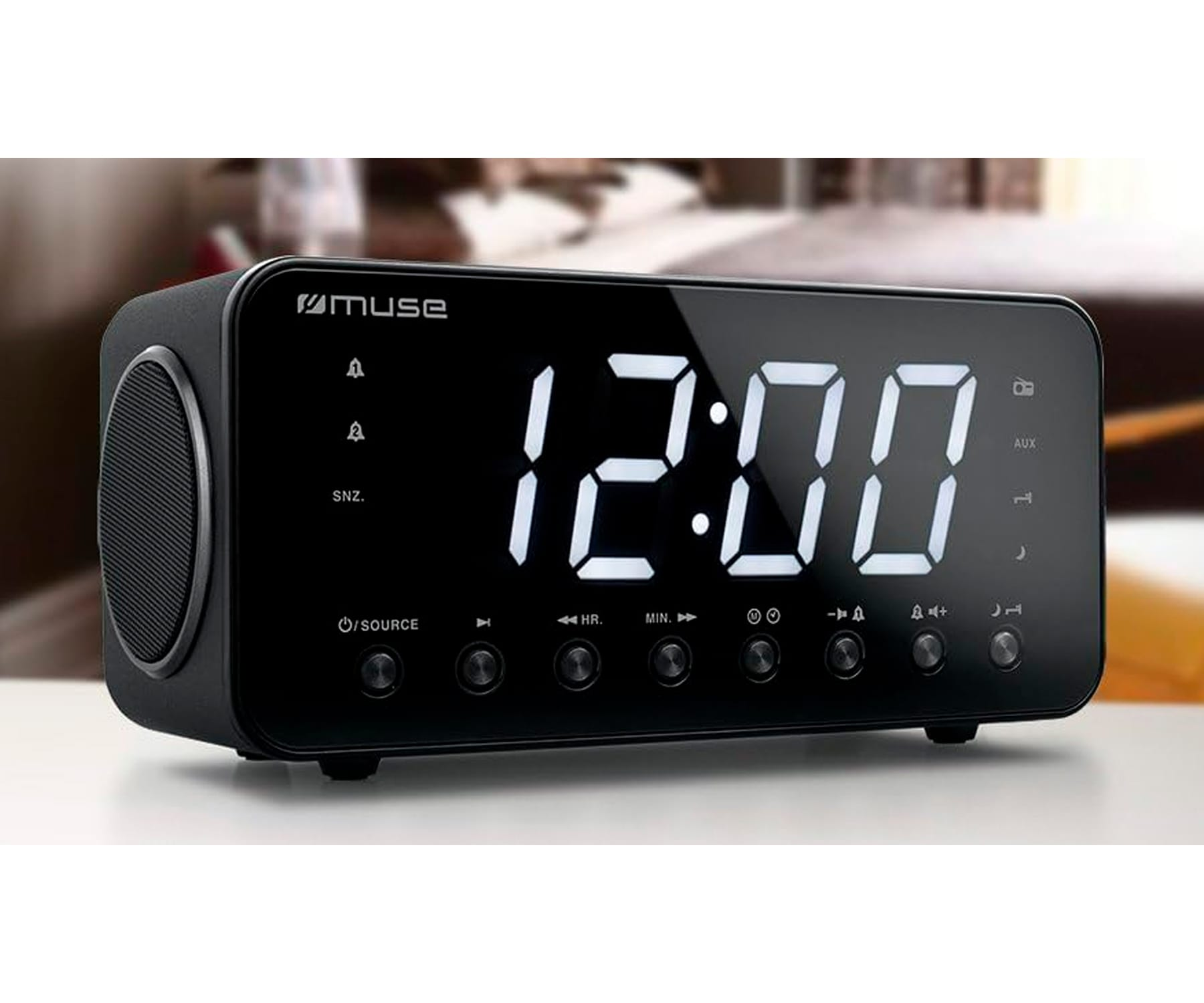 Czarny budzik cyfrowy z godziną 12:00. Umieszczony na białej powierzchni. Rozmyte tło.