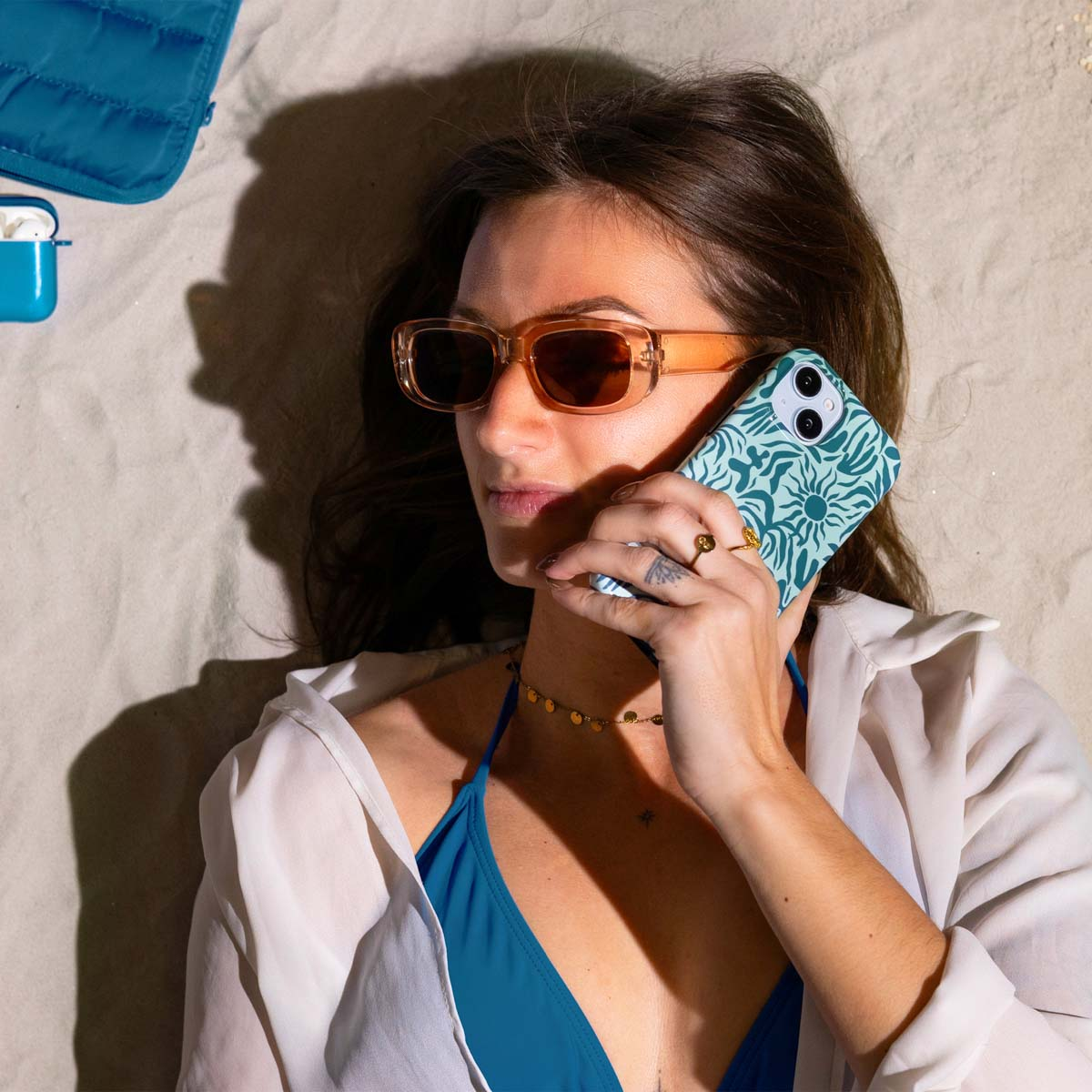 Vrouw op strand met zonnebril, telefoon. Blauwe bikini en tas.