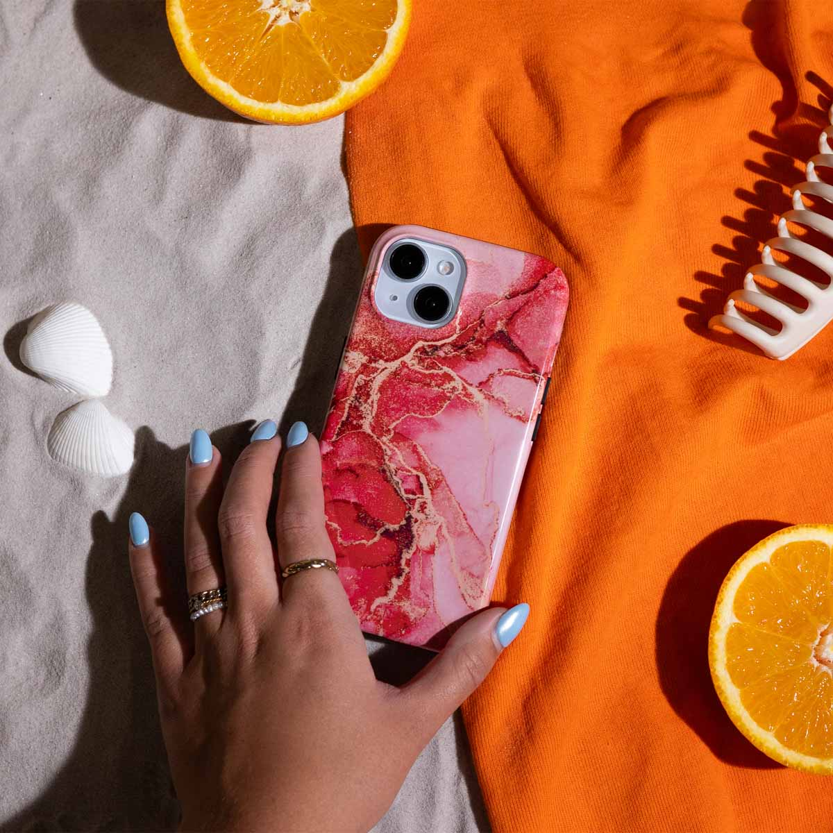 Main avec ongles bleus tenant une coque de téléphone avec motif marbre rose, à côté d'orange et coquillages.