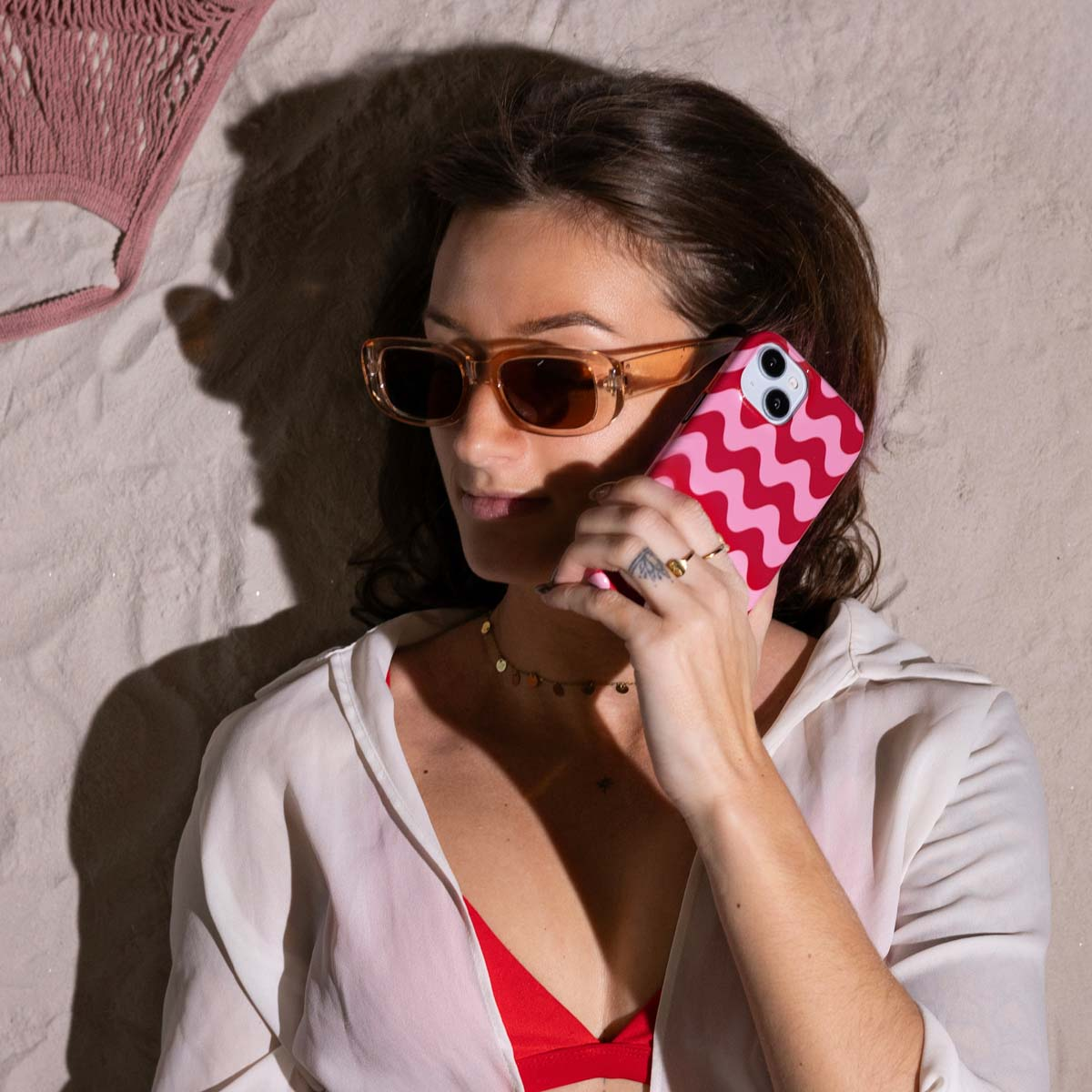 Een vrouw met zonnebril houdt een telefoon vast in een roze hoesje met patroon. Ze is op het strand.