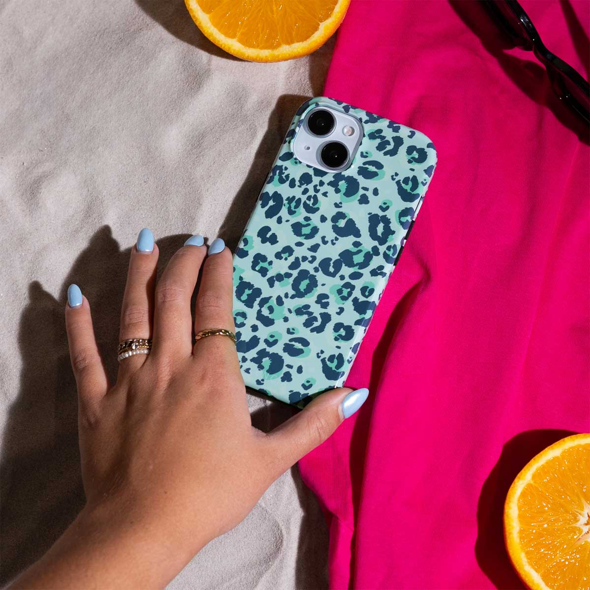 Hand met blauwe nagels houdt teal leopard print telefoonhoes vast. Sinaasappelschijf en roze shirt achtergrond.