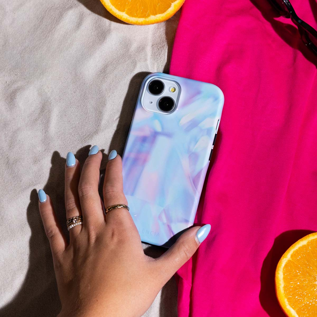 Main avec ongles bleus tenant une coque de téléphone avec un motif marbre bleu, à côté de tissu rose et orange.