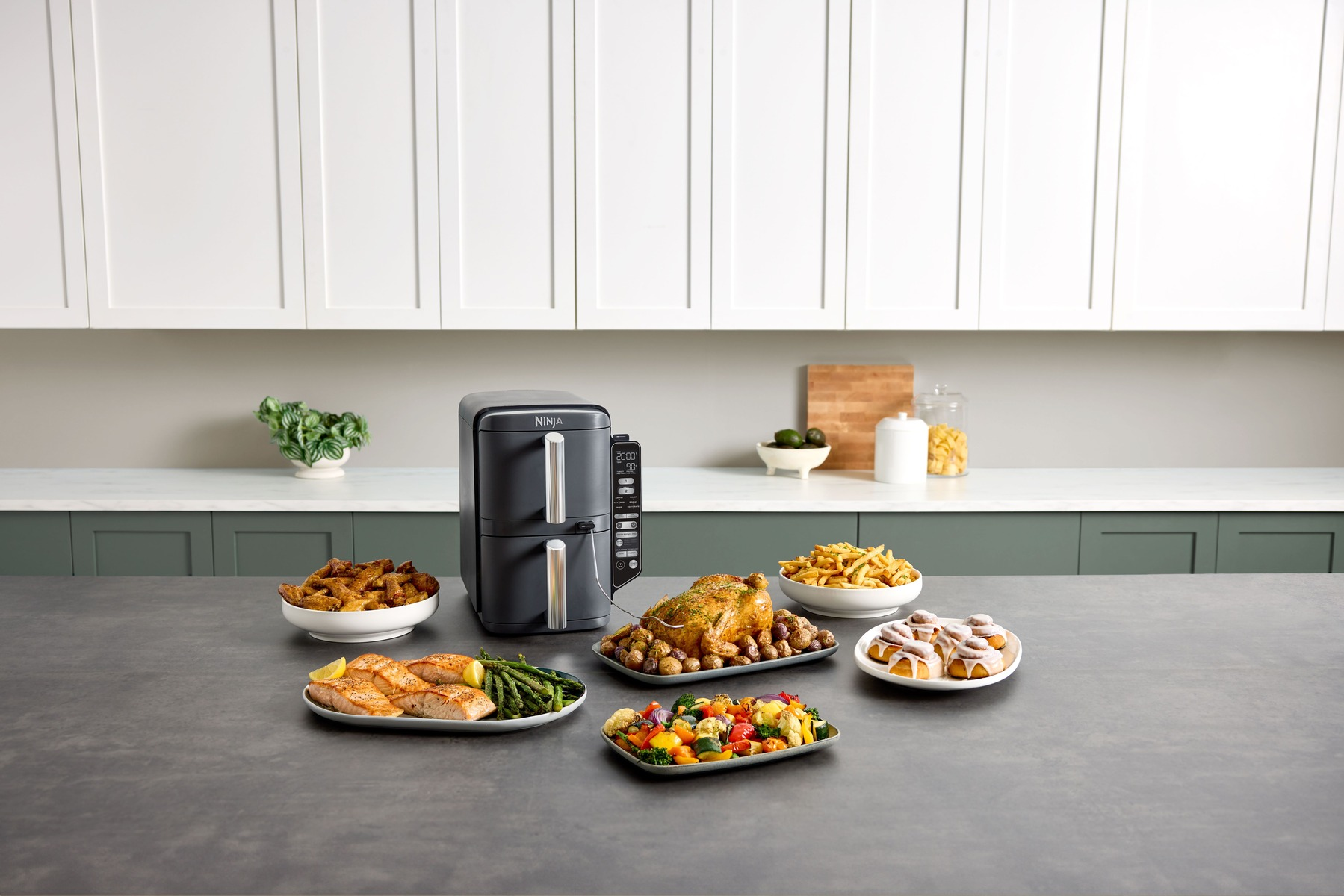 Frytownica powietrzna z przygotowanym jedzeniem: kurczak, łosoś, frytki i bułki na blacie.