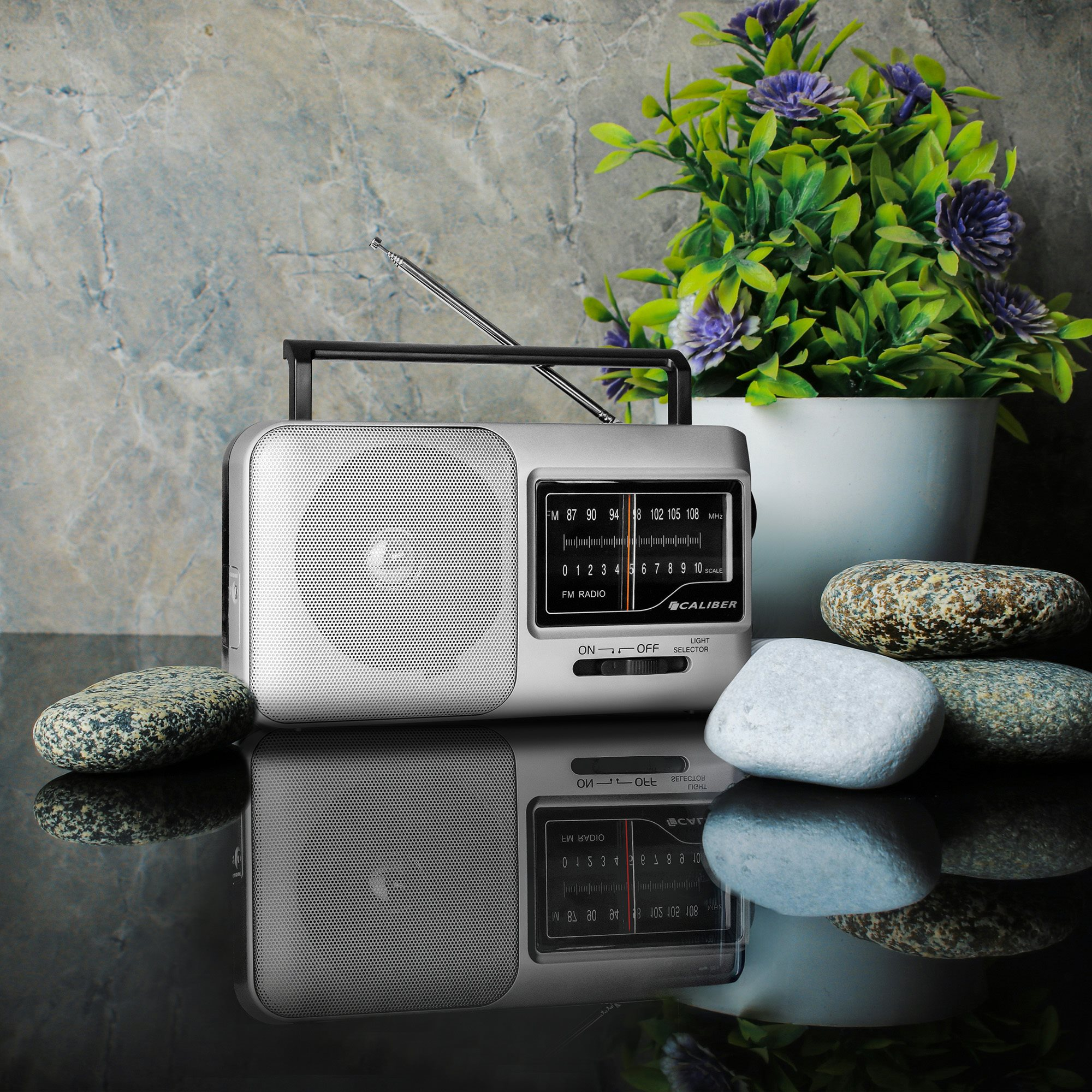Silver radio with a black handle, and a small plant and stones on a reflective surface.