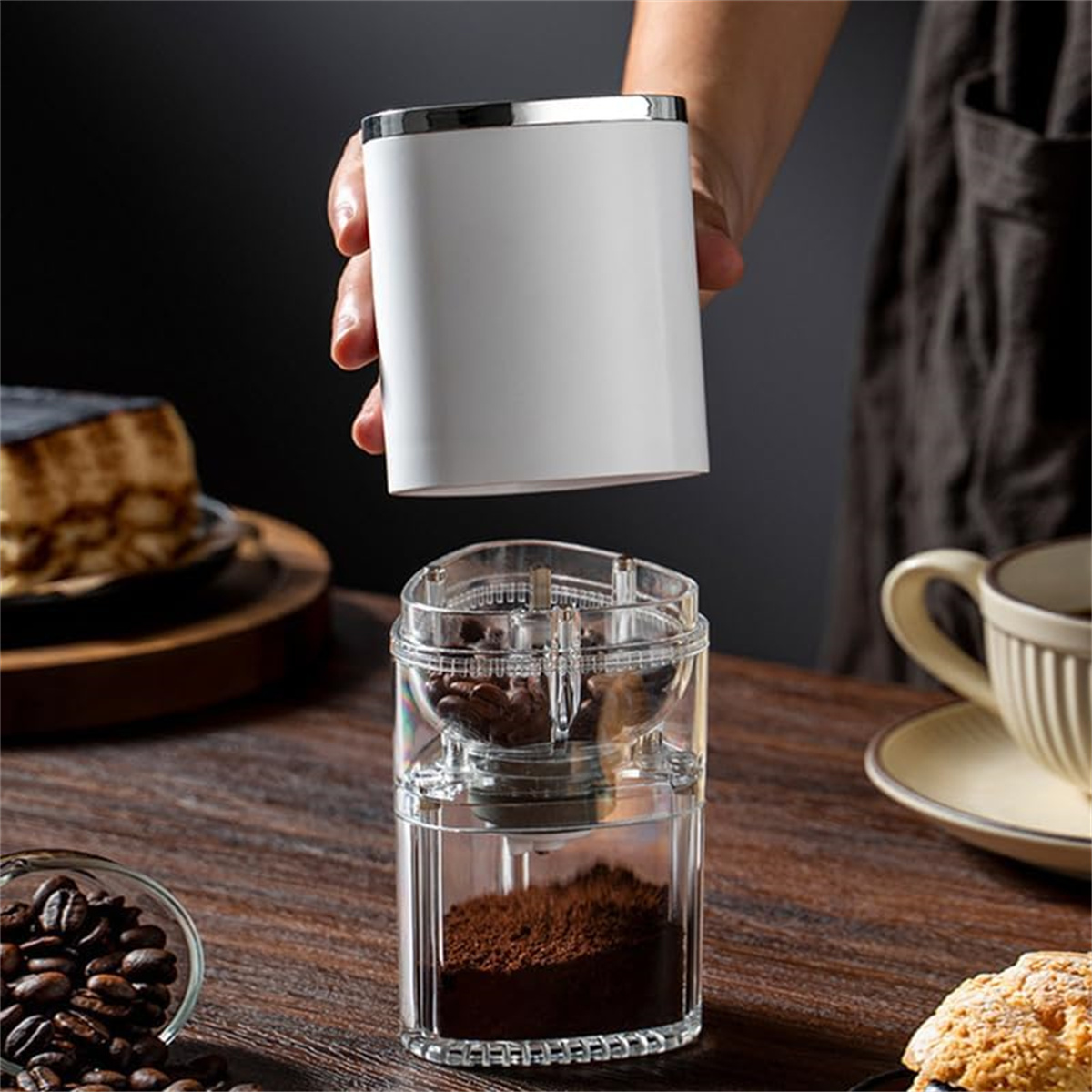 Eine Person hält eine weiße Kaffeemühle, Kaffeebohnen und gemahlenen Kaffee auf einem Holztisch.