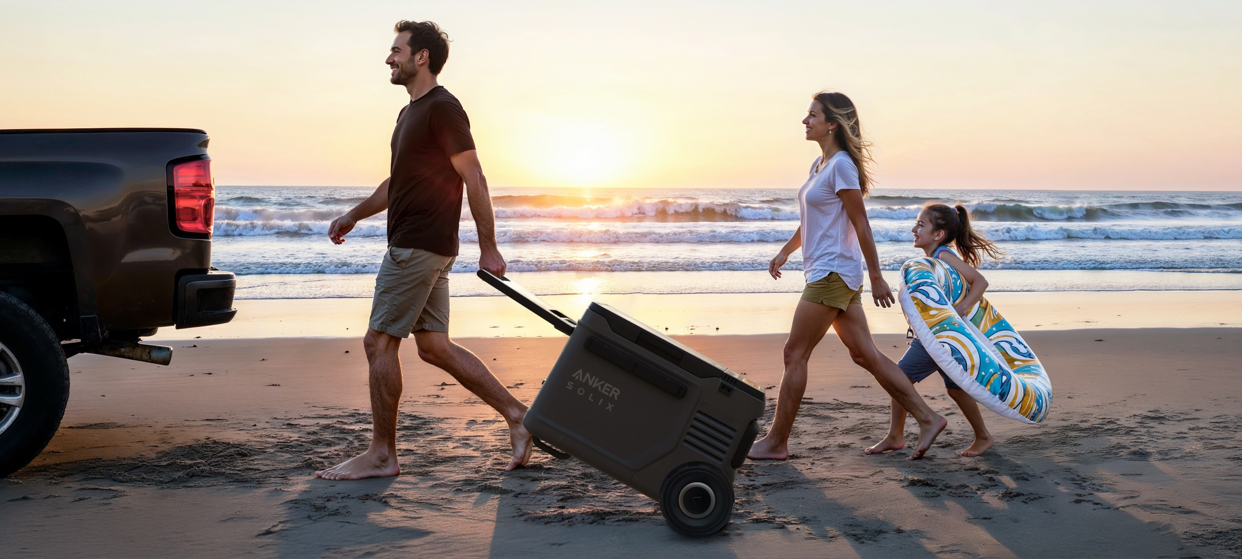 Rodzina idąca po plaży, ciągnąca chłodnicę na kółkach. Zachód słońca w tle.