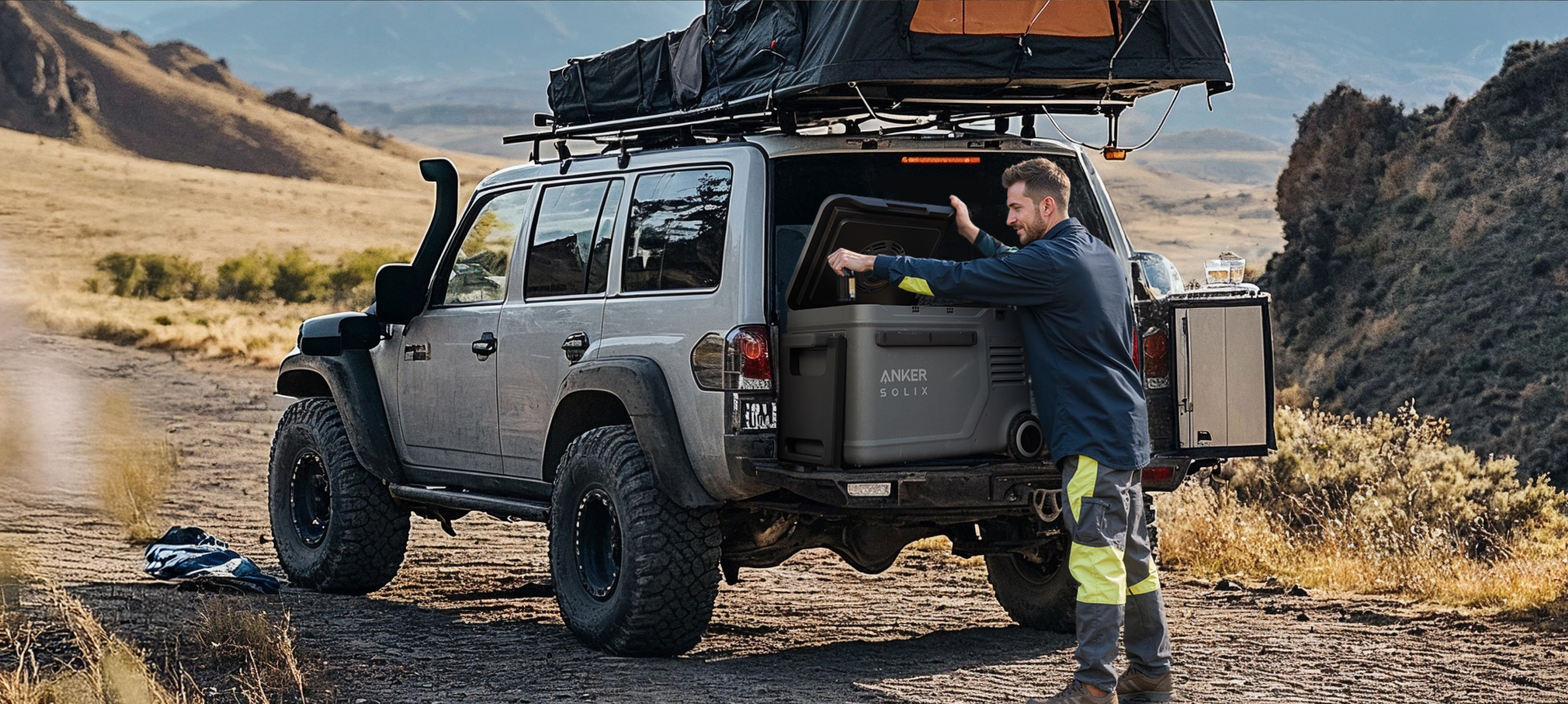 Mężczyzna ładuje chłodziarkę do tyłu szarego SUV-a na polnej drodze. Namiot jest na dachu.