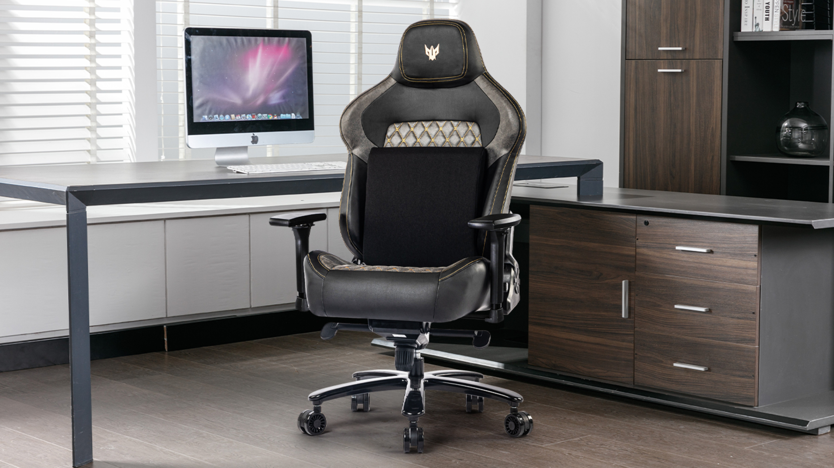 Schwarz-grauer Gaming-Stuhl. Schreibtisch und Computer im Hintergrund, Fenster dahinter. Holzschrank rechts.