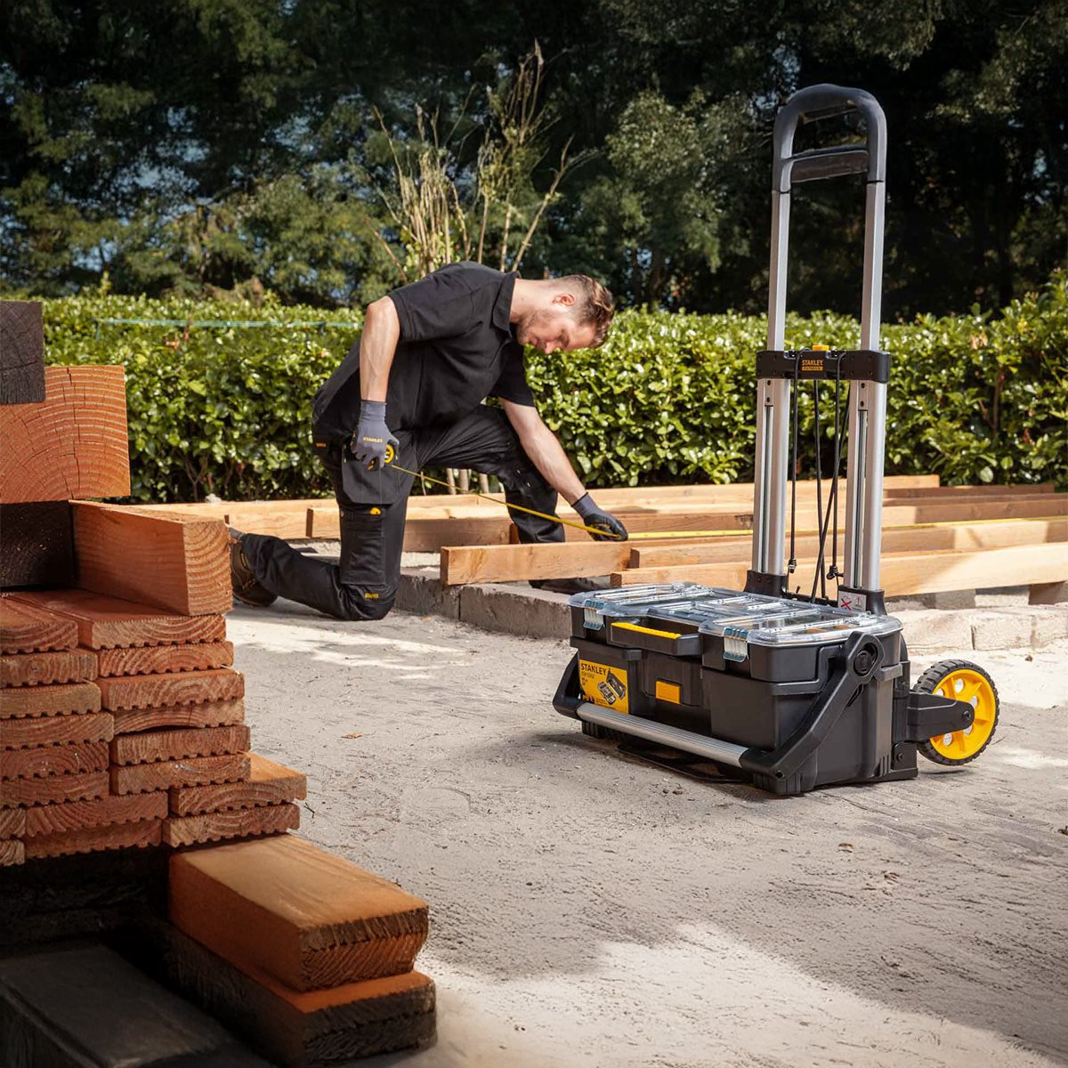 Man kneeling, measuring wood on a construction site. A cart holds tools and supplies.