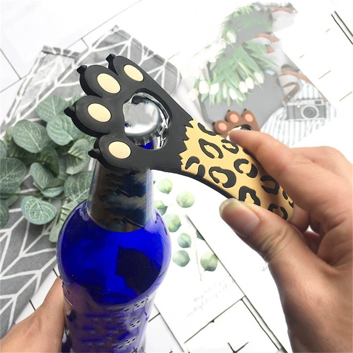 Flaschenöffner mit Leopardenmuster in der Hand, öffnet eine blaue Flasche. Weißer Hintergrund.