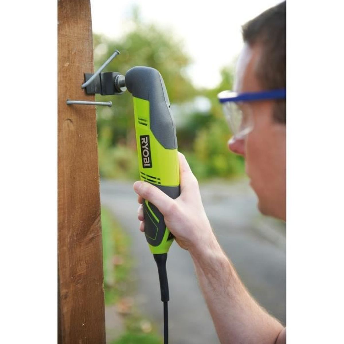 Ein Mann benutzt ein grün-schwarzes Ryobi-Elektrowerkzeug auf Holz. Blaue Schutzbrille wird getragen.