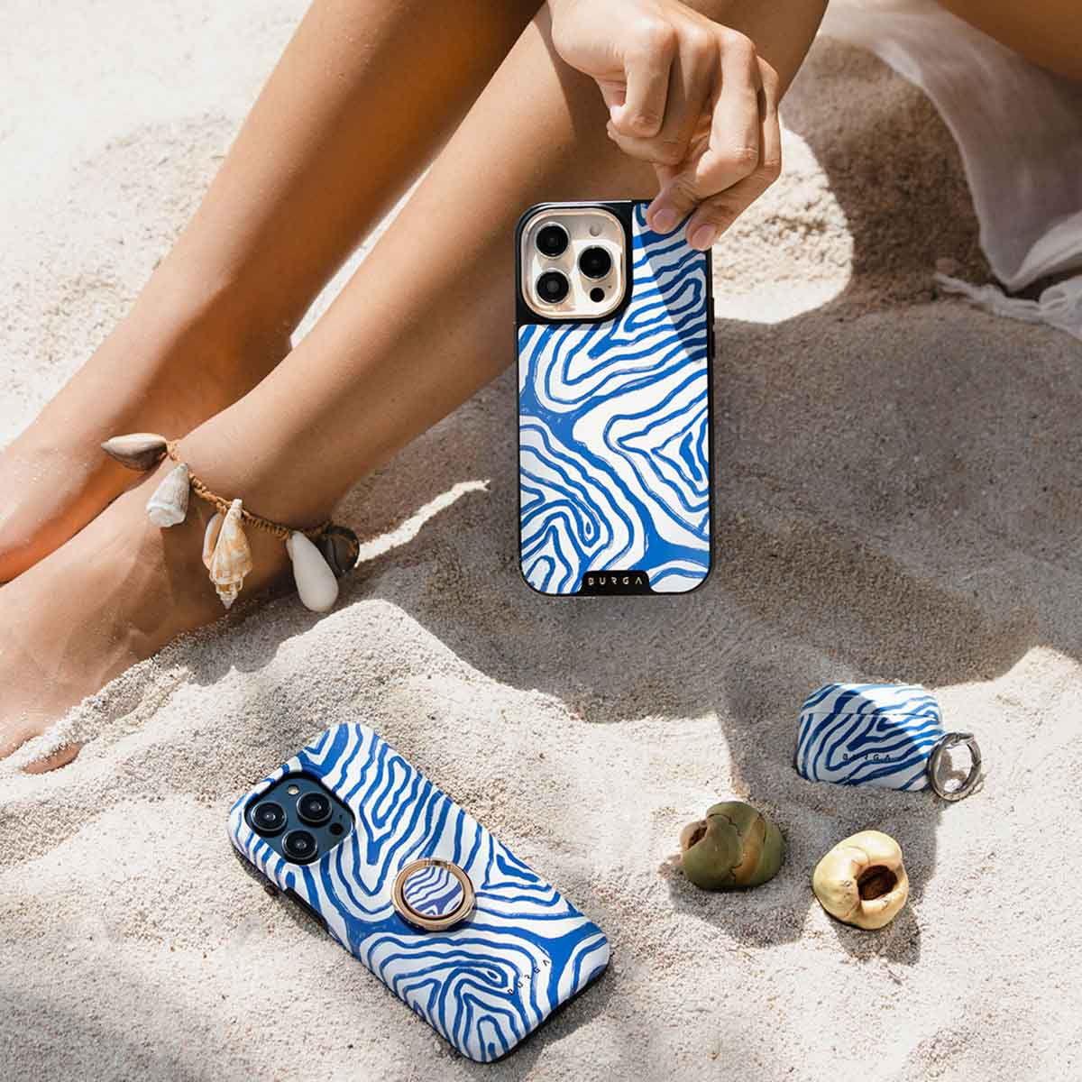 Benen van een persoon met een schelpenenkelband, die een telefoon met een blauw-witte hoes op het zand vasthoudt.