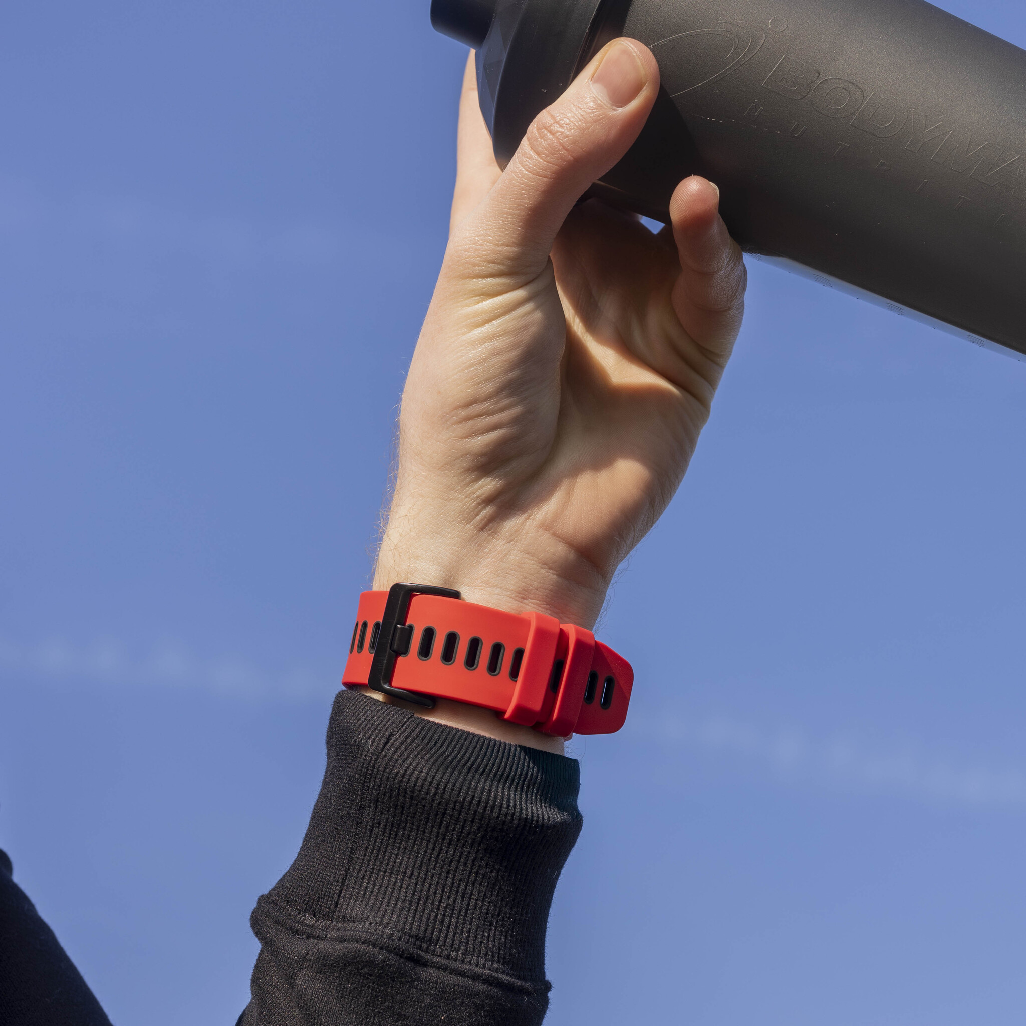 Der Arm einer Person mit einem roten Fitnessband. Sie halten eine schwarze Flasche, mit blauem Himmel im Hintergrund.