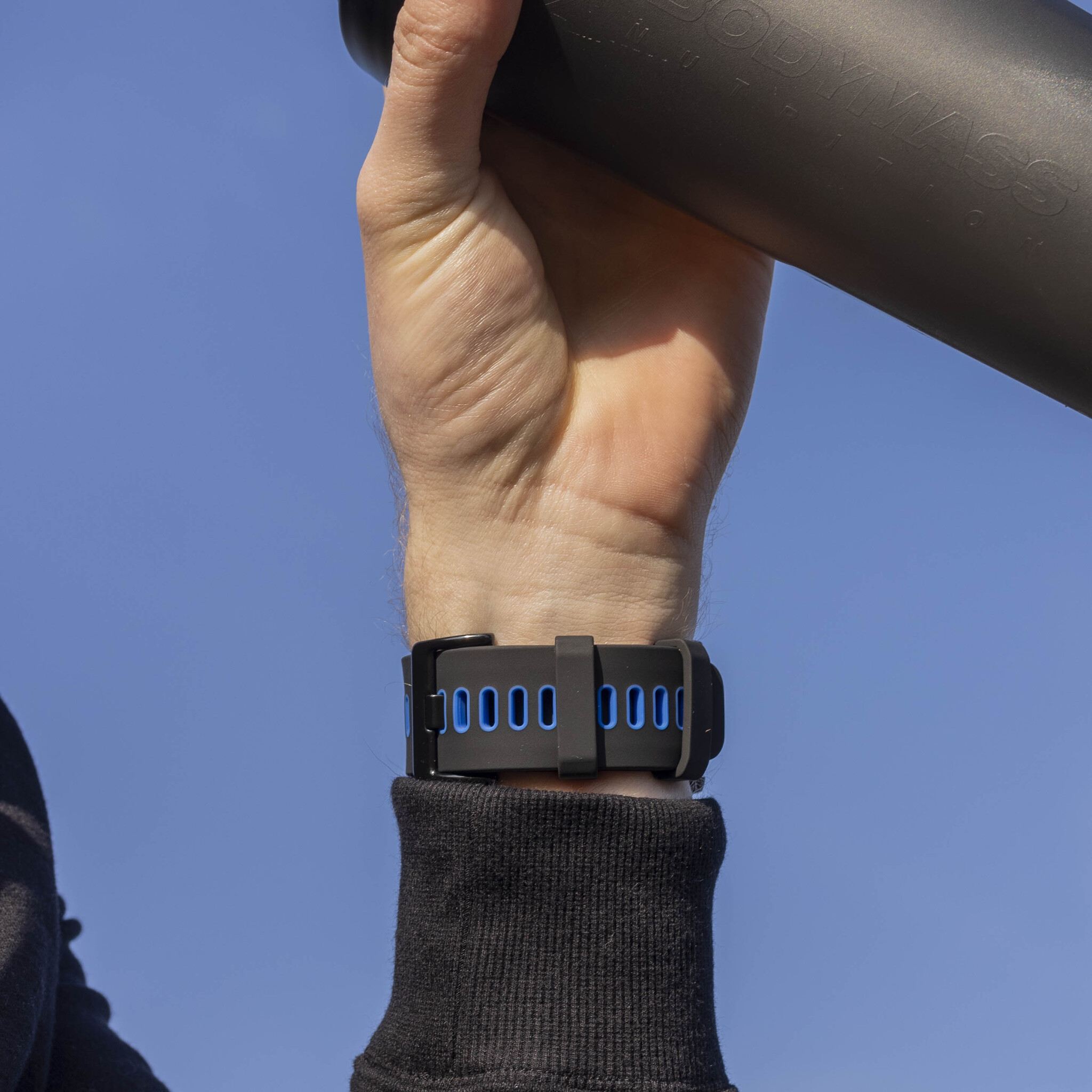 Handgelenk mit schwarzer Uhr und blauen Akzenten, hält eine schwarze Flasche, blauer Himmel.