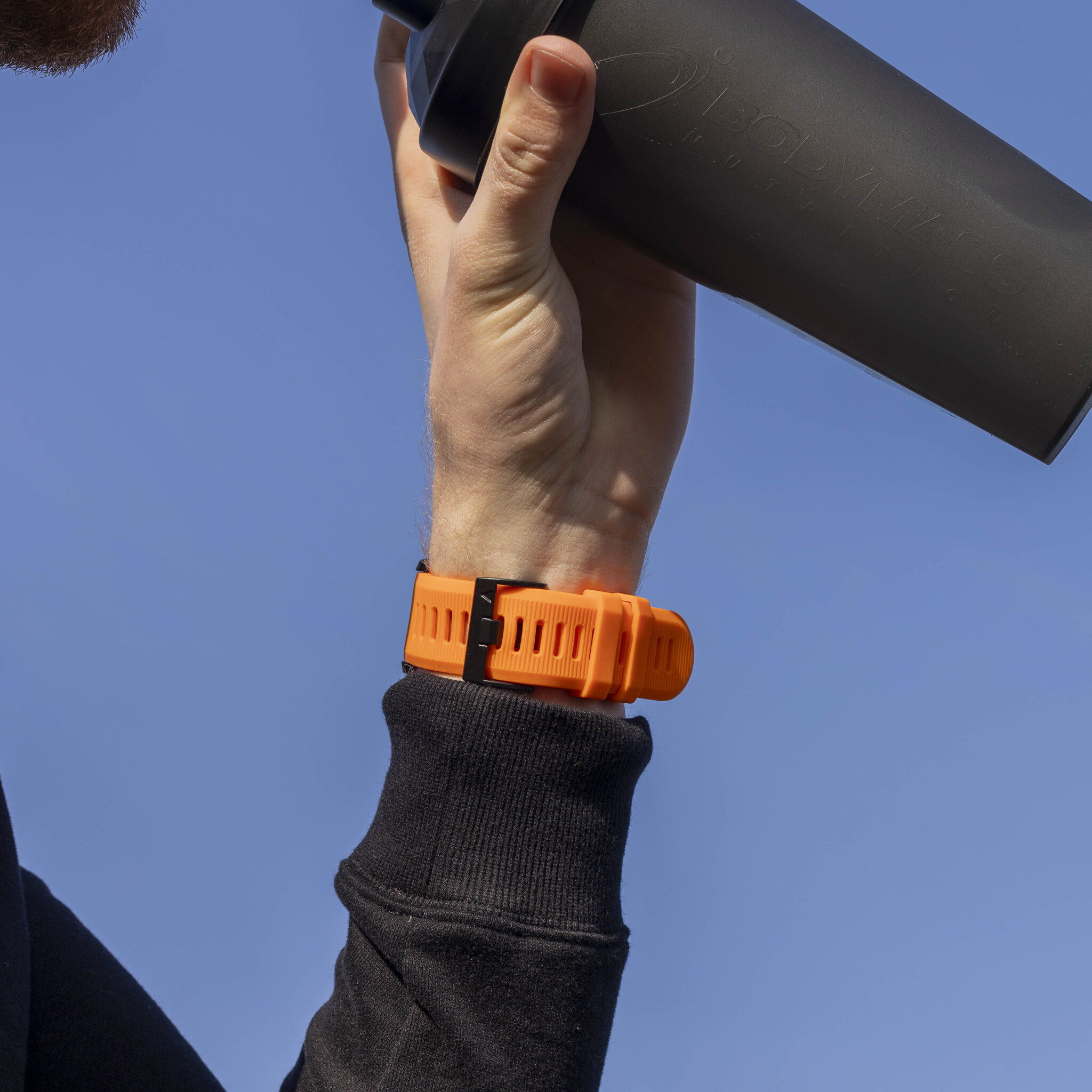 Eine Hand, die eine schwarze Shaker-Flasche hält und einen orangefarbenen Fitnesstracker trägt.