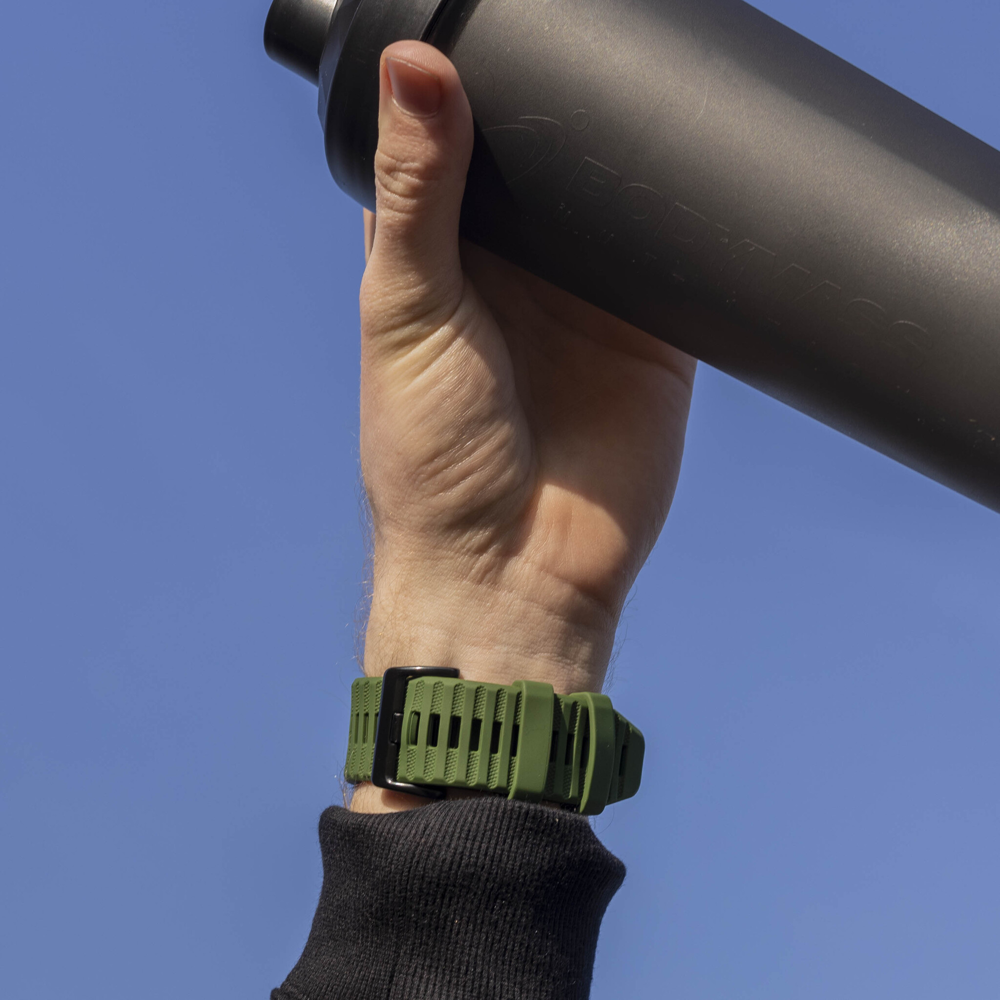 Eine Hand hält eine schwarze Wasserflasche mit einem grünen Armband. Blauer Himmel im Hintergrund.