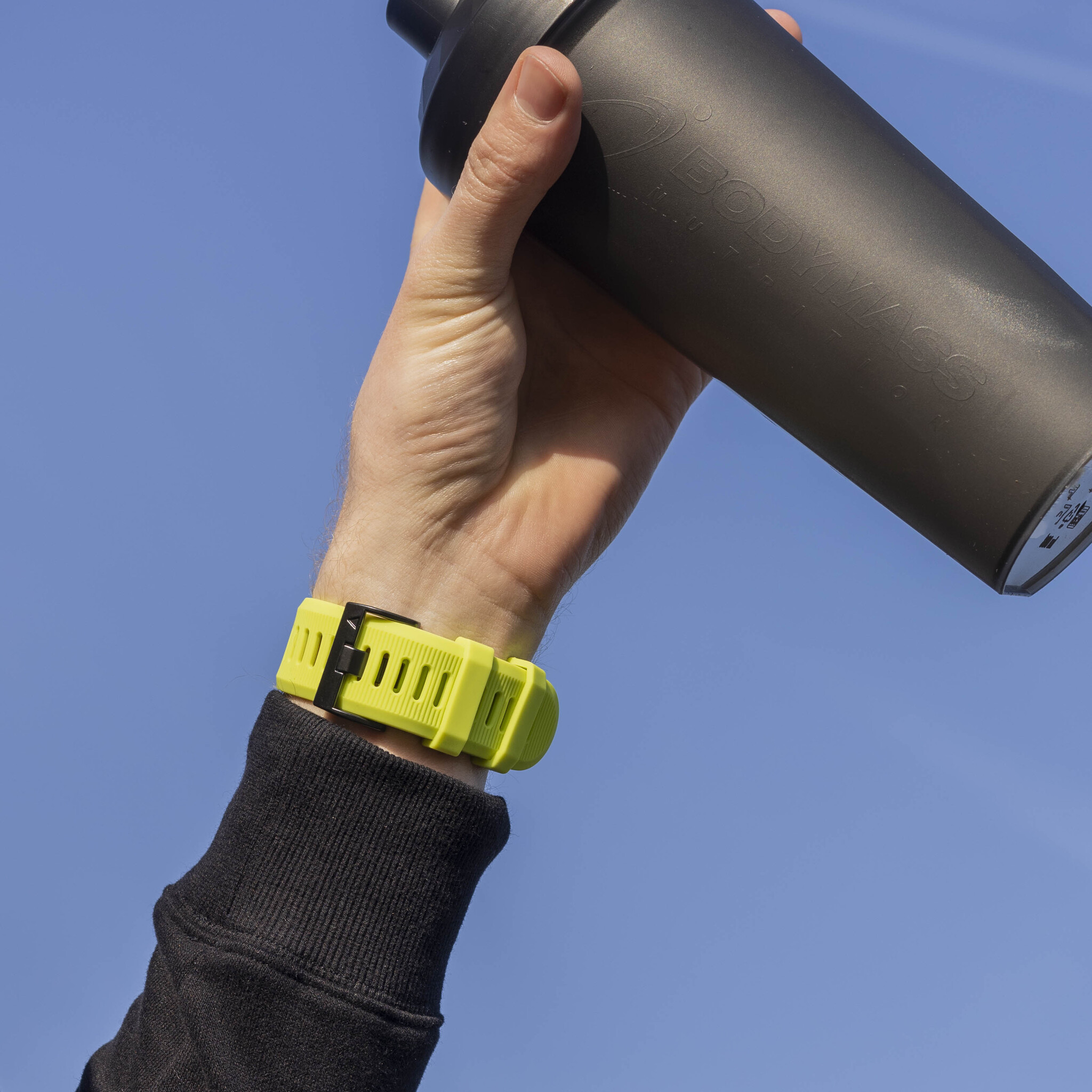 Eine Person hält eine schwarze Shaker-Flasche. Ein Handgelenk mit gelbem Armband ist sichtbar.