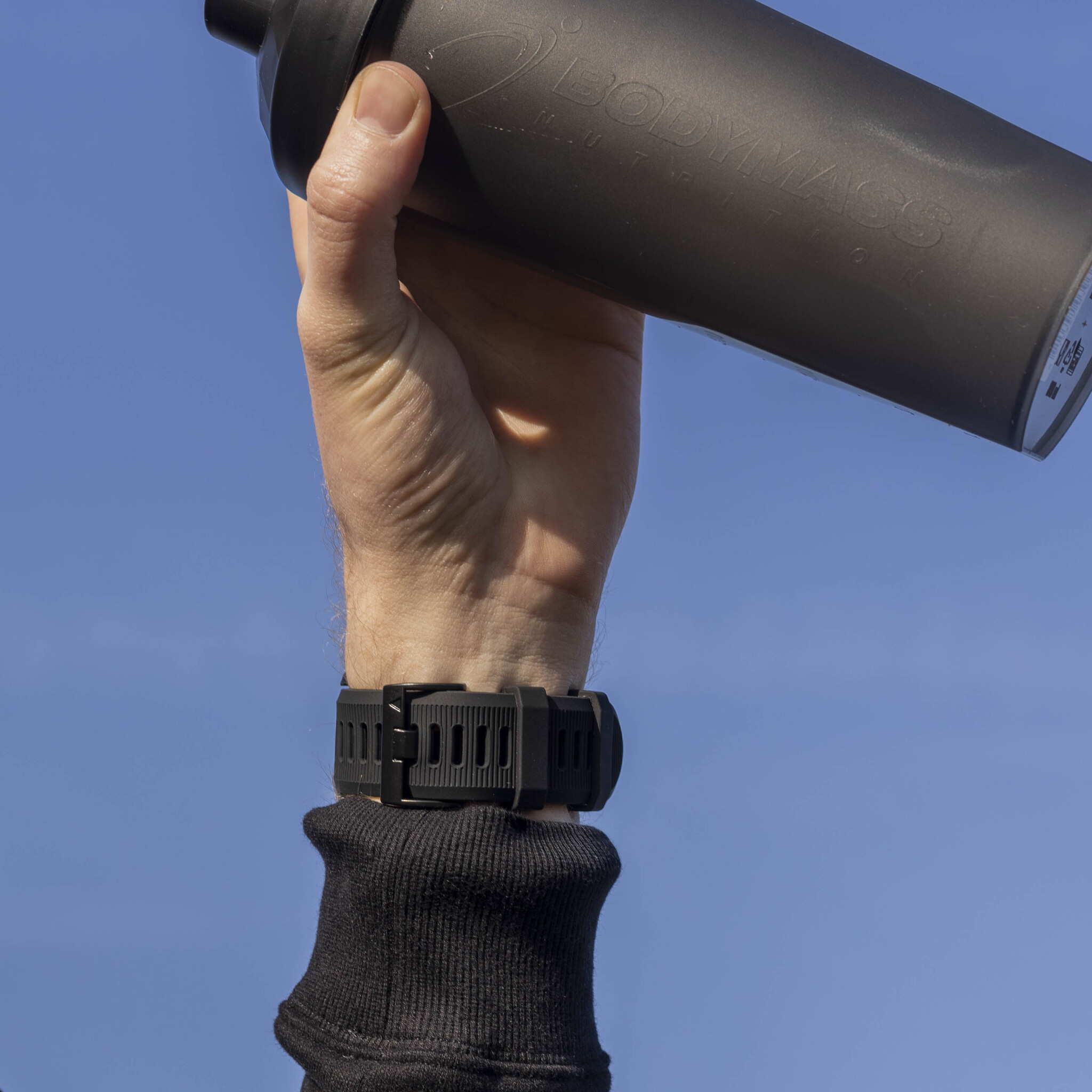 Die Hand einer Person, die eine schwarze Shaker-Flasche vor blauem Himmel hält.