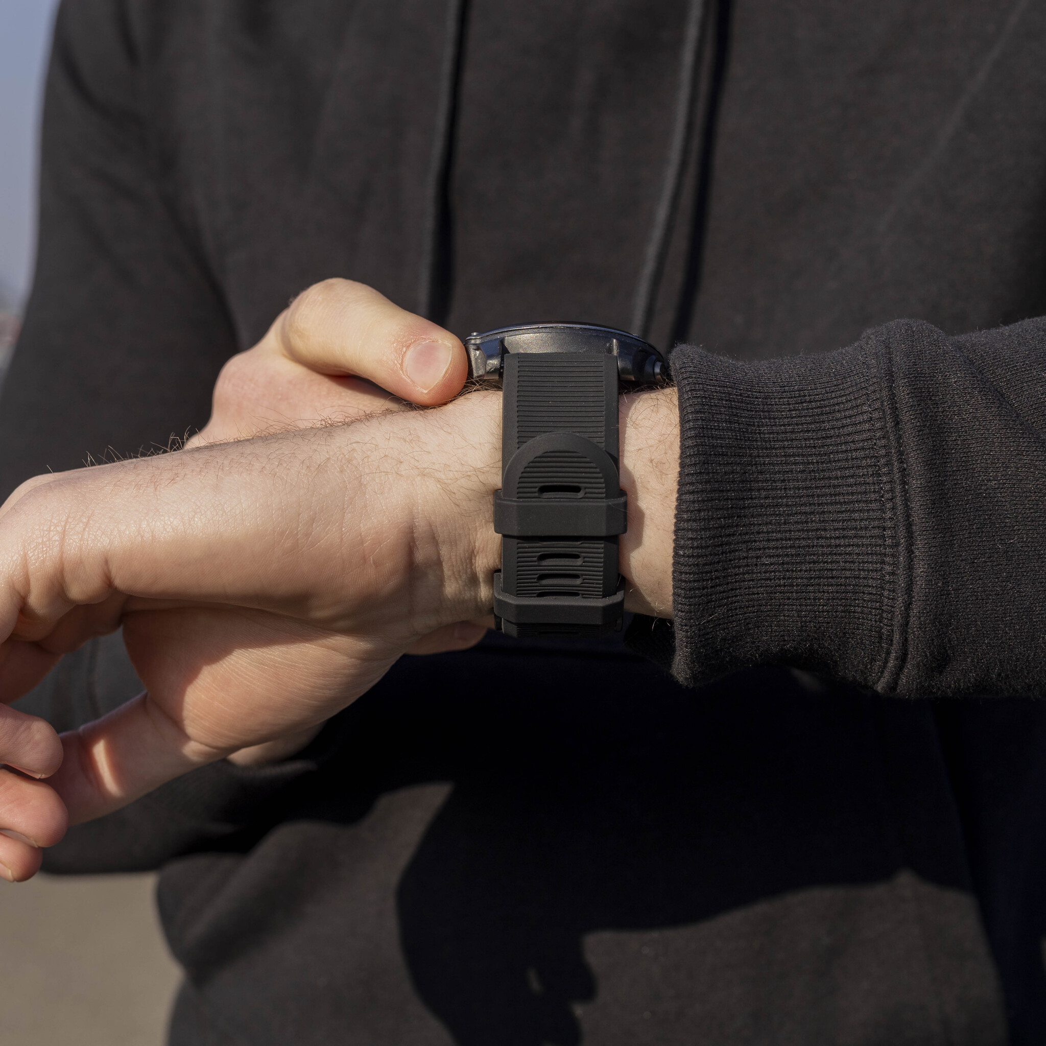 Hand mit schwarzer Uhr. Person trägt schwarzen Kapuzenpullover. Der Hintergrund ist unscharf.