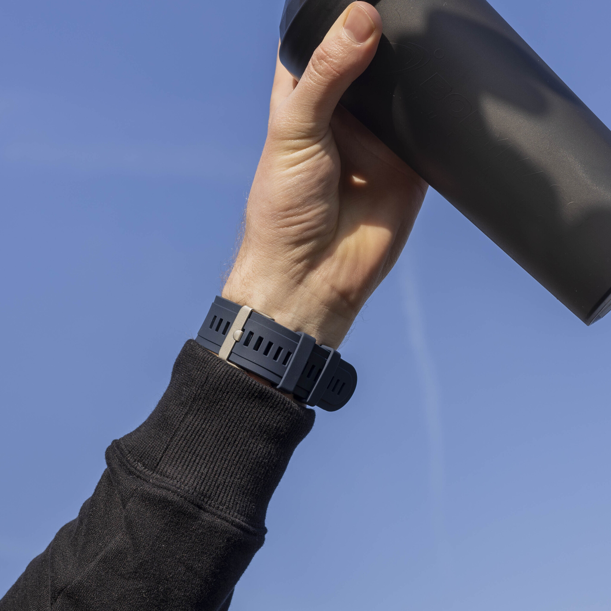 Eine Hand, die eine schwarze Shaker-Flasche vor blauem Himmel hält.