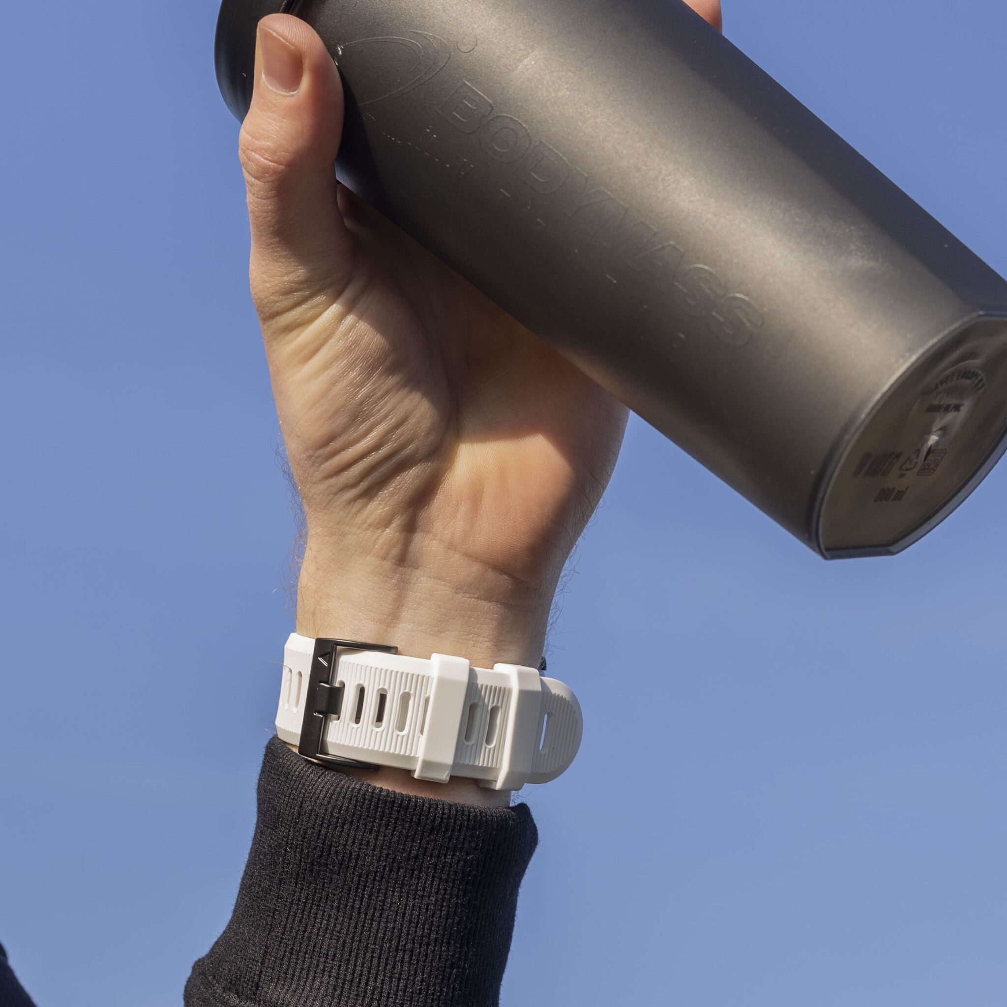 Arm hält schwarze Shakerflasche mit weißer Uhr, vor blauem Himmel.