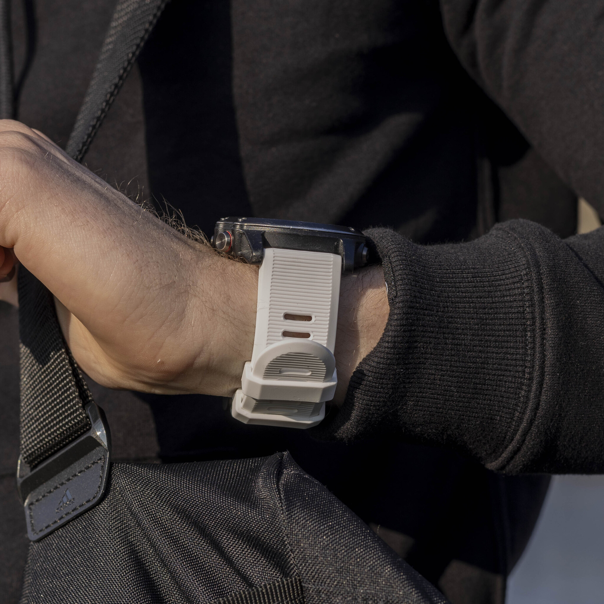 Handgelenk mit Uhr und weißem Armband. Schwarze Tasche mit Riemen an der Seite.