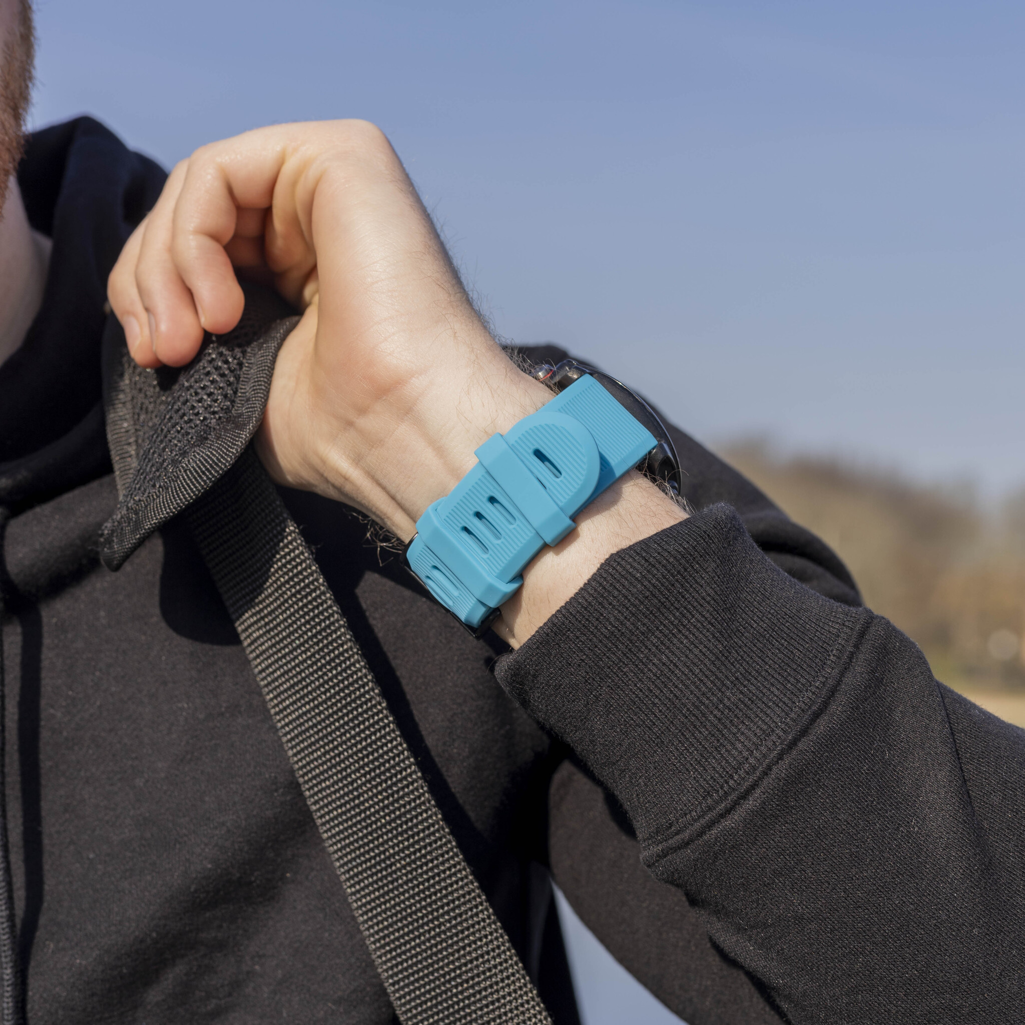 Handgelenk mit blauem Uhrenarmband. Person trägt schwarzen Kapuzenpullover und hält eine Tasche.