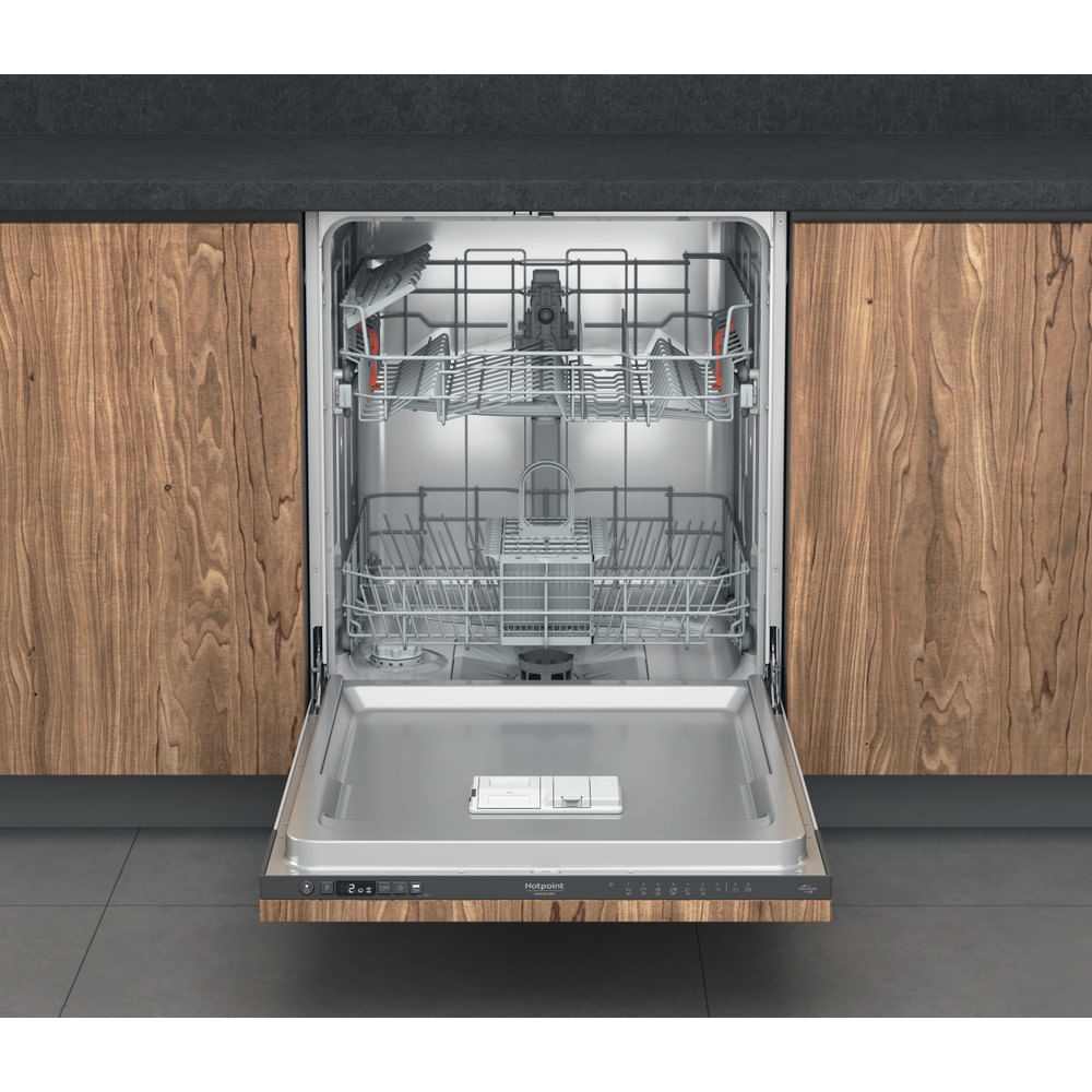 Lavastoviglie Hotpoint aperta che mostra le griglie interne, con uno sfondo di mobili in legno e ripiani grigi.