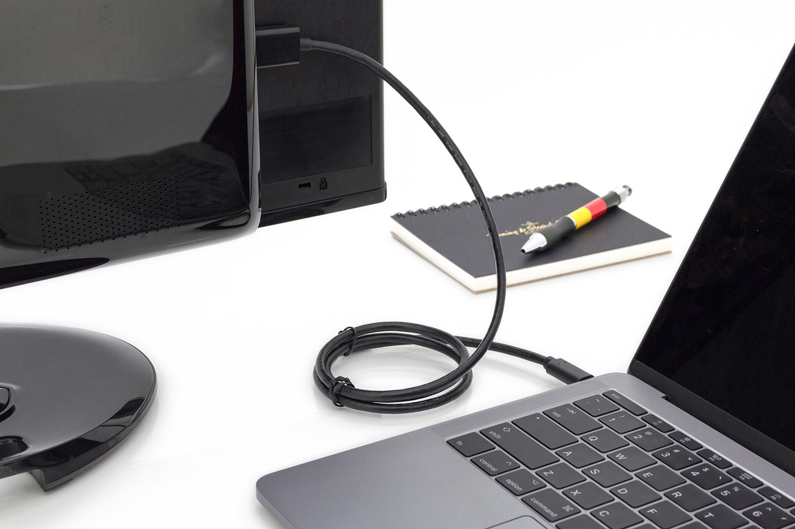 Ein Laptop ist mit einem Monitor verbunden. Ein schwarzes Kabel ist angeschlossen. Notizbuch und Stift.