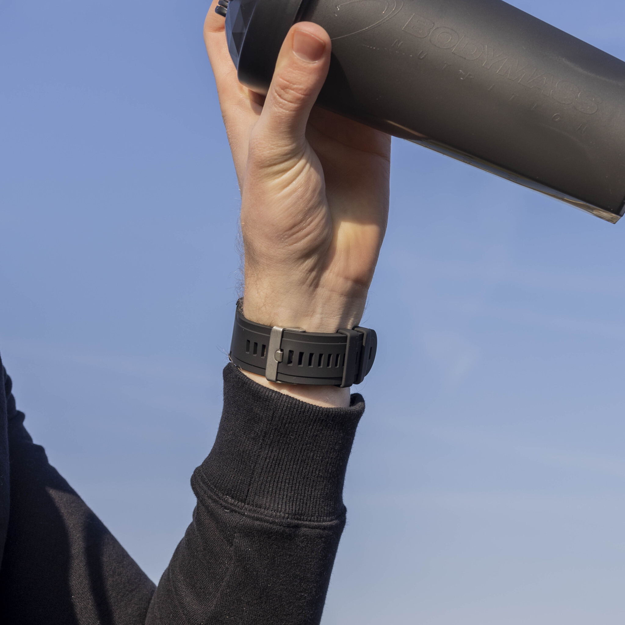 Hand, die einen schwarzen Shaker hält, mit einer Uhr am Handgelenk. Der Himmel ist blau.