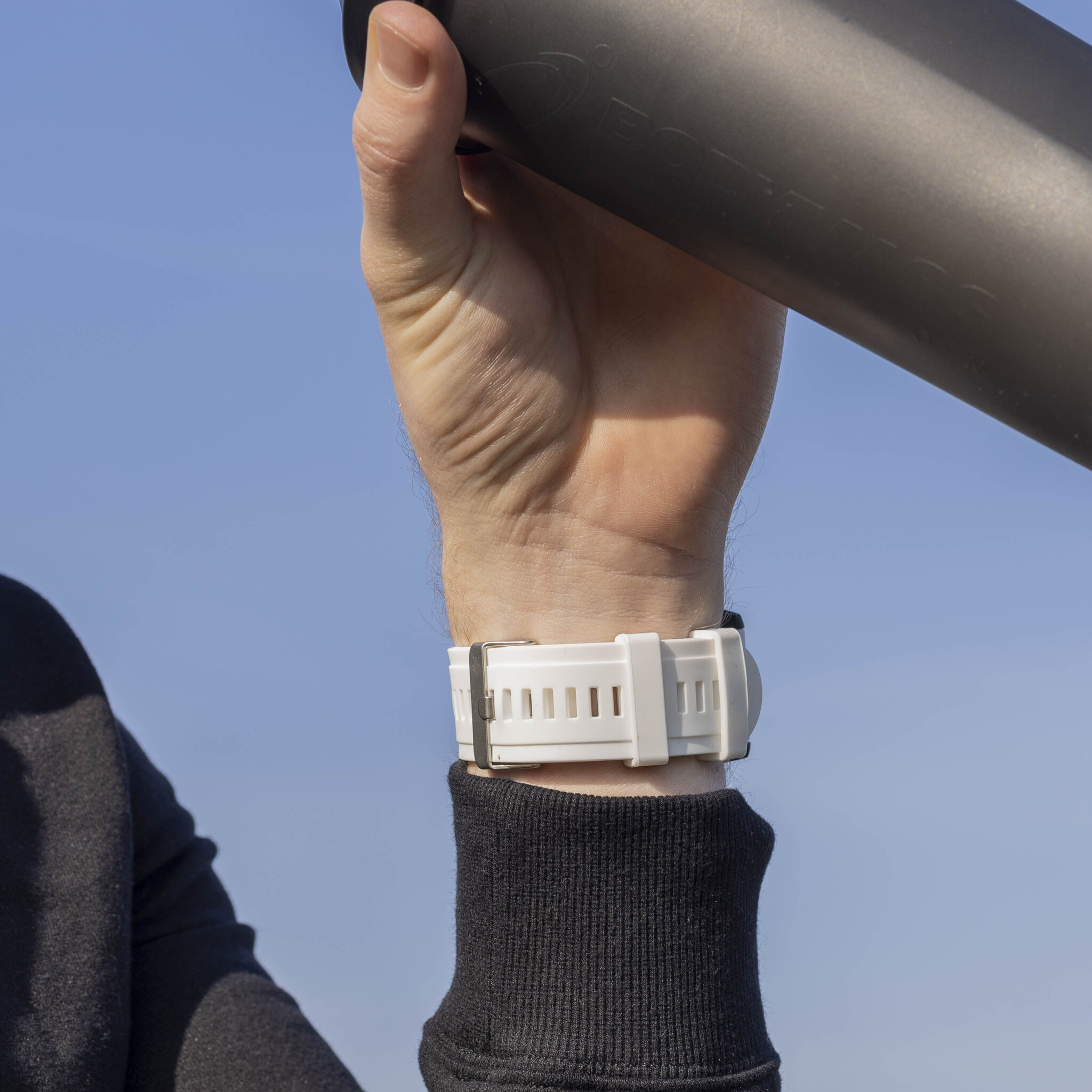 Ein Handgelenk mit einer Uhr mit weißem Armband. Die Person trägt eine schwarze Jacke.