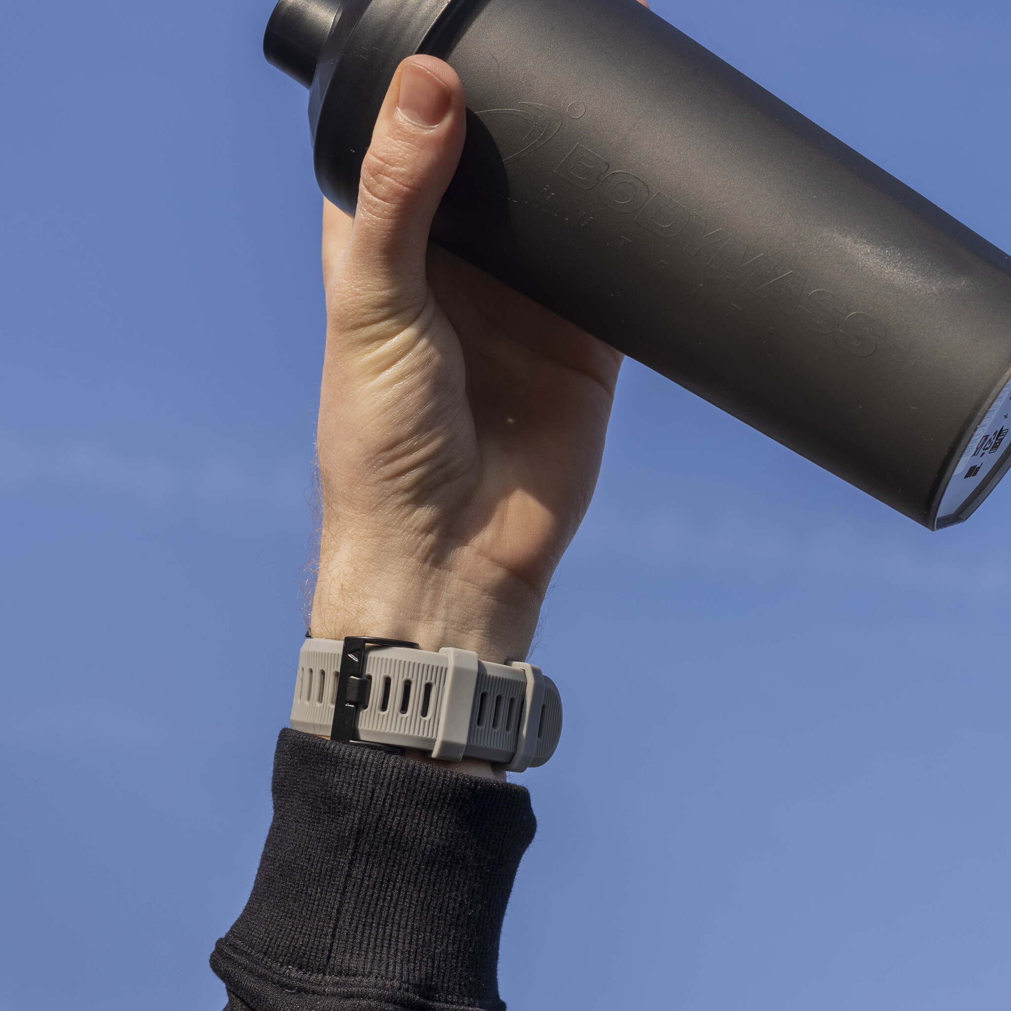 Hand hält eine schwarze Shaker-Flasche vor blauem Himmel, mit Uhr am Handgelenk.