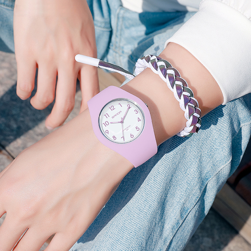 Das Handgelenk einer Person mit einer rosa Uhr und einem geflochtenen Armband in Weiß und Lila.