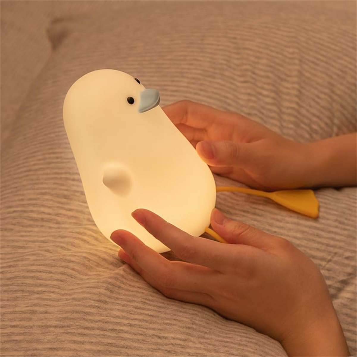 Ein weißes pinguinförmiges Nachtlicht in Händen, mit gelbem Schwanz, auf einem Bett.