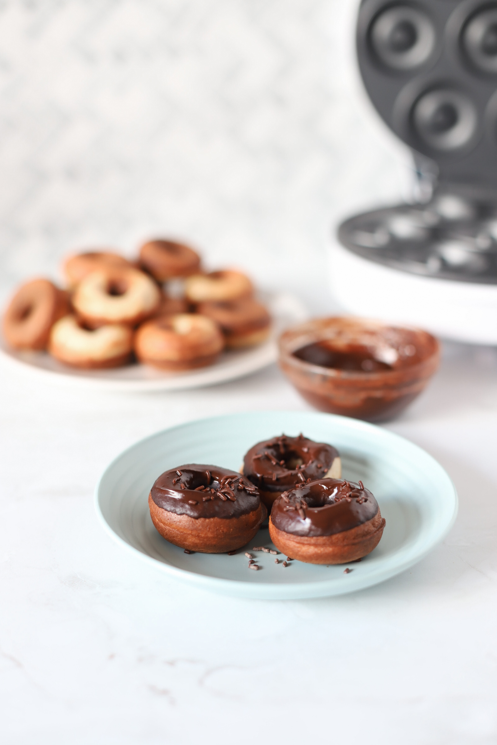 Donuts en plato con glaseado de chocolate. Máquina de donuts al fondo.