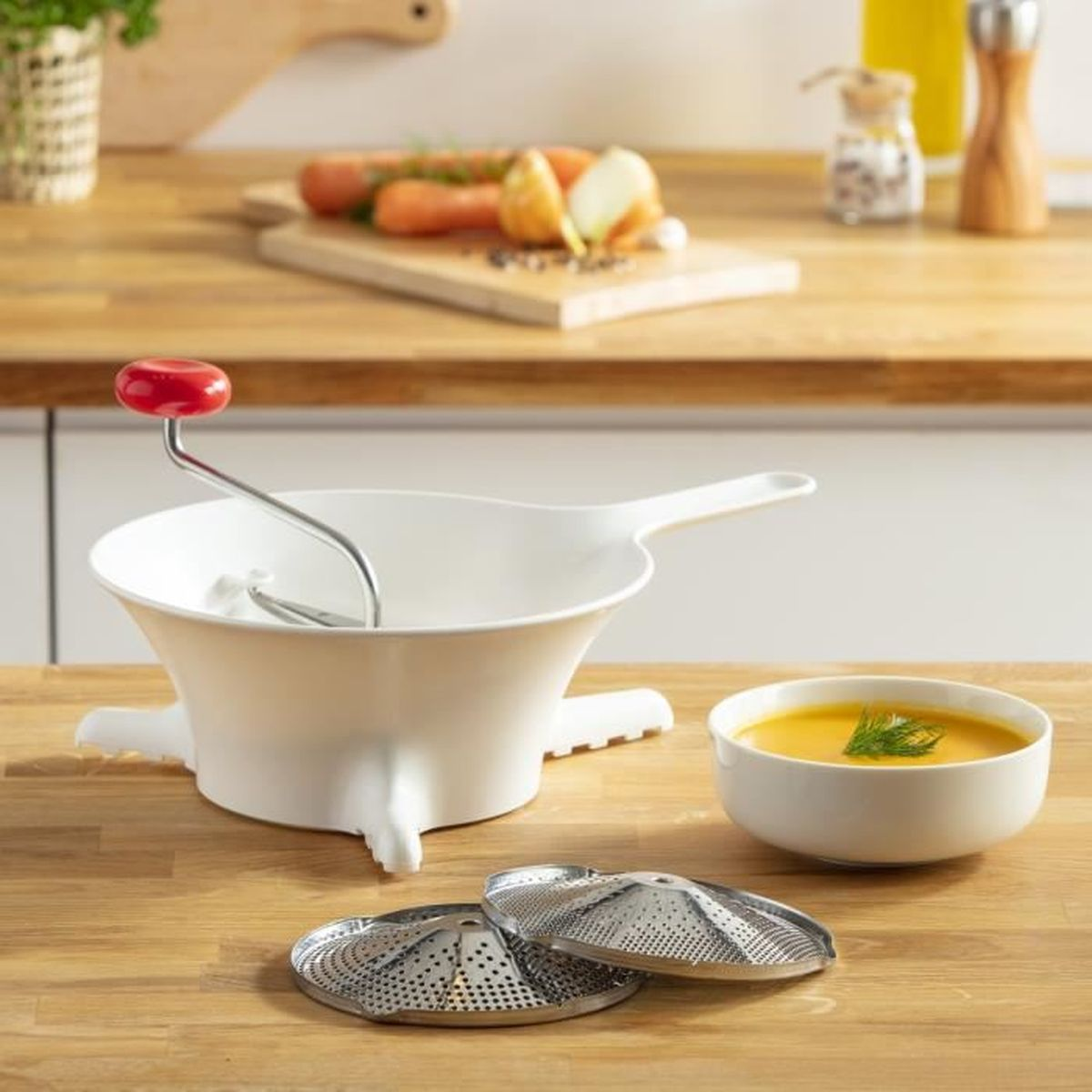 Passaverdure bianco con manico rosso, due colini e una ciotola di zuppa su superficie in legno.