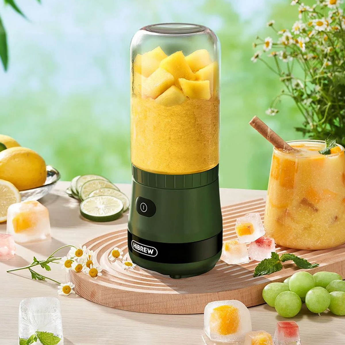 Zielony przenośny blender. Zawiera żółte owoce. W tle kostki lodu, plasterki owoców i napój.