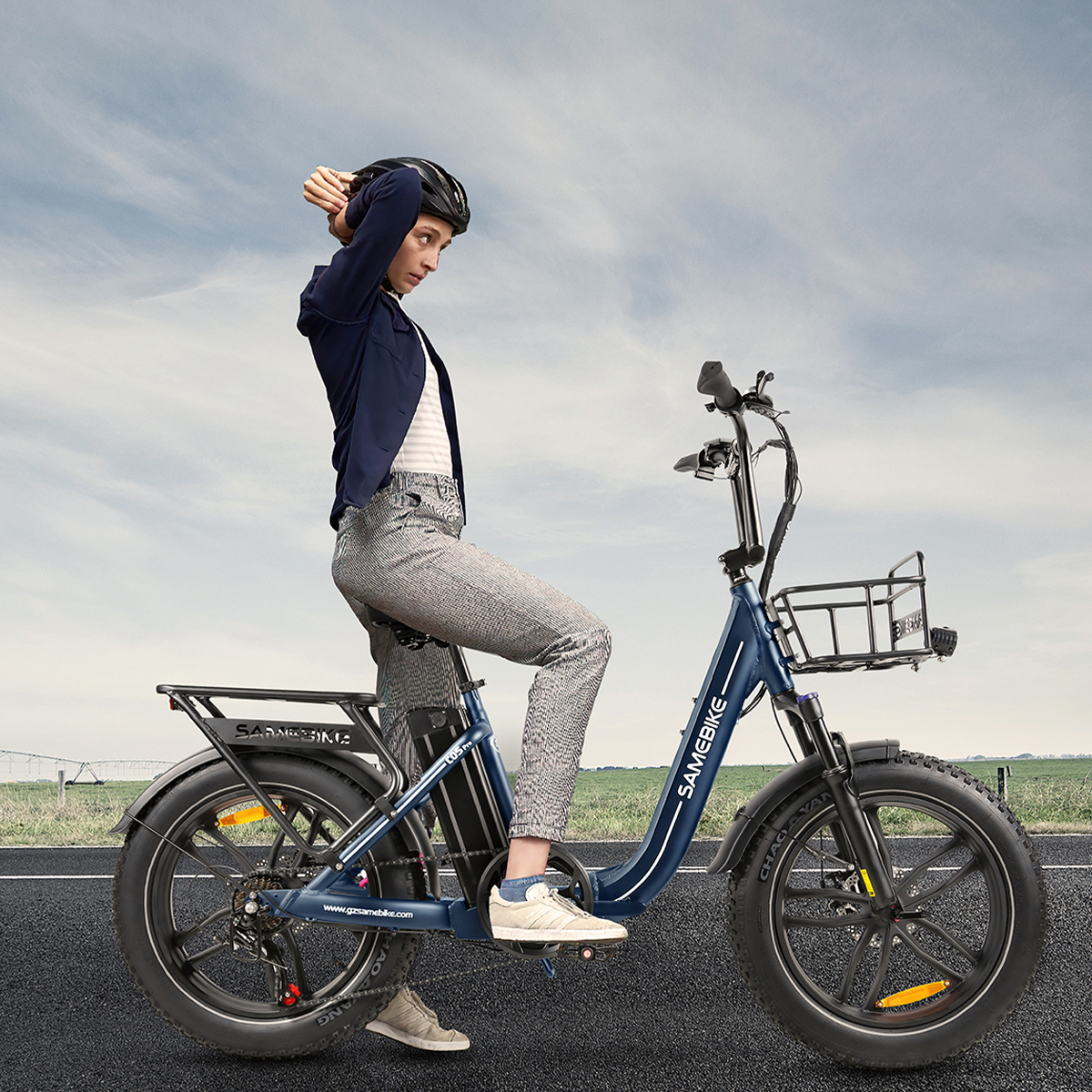 Kobieta na niebieskim rowerze elektrycznym na drodze, błękitne niebo. Rower ma kosz, logo Samisbike.