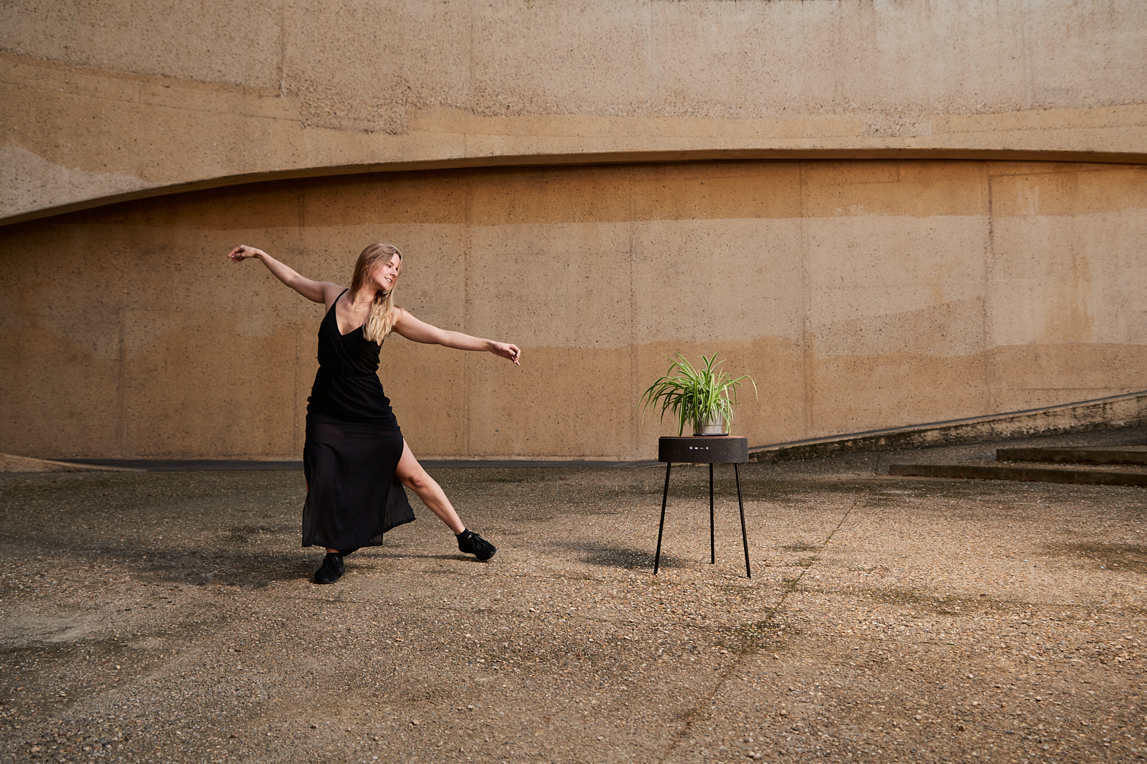 Een vrouw in een zwarte jurk danst voor een betonnen muur en een plant.