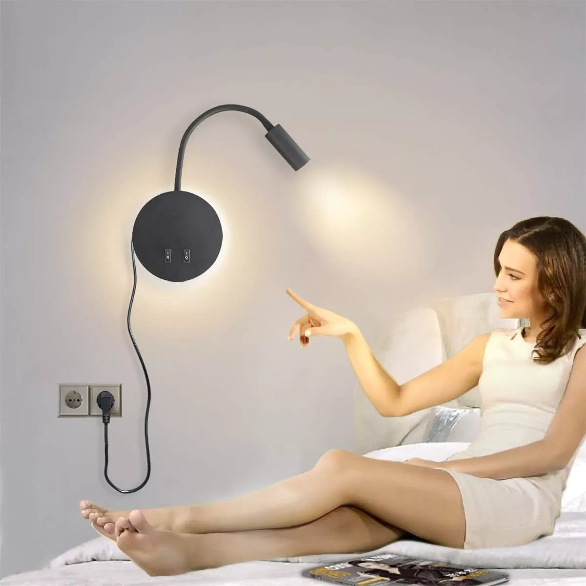 Frau zeigt auf eine Wandlampe. Die Wandlampe hat einen schwarzen Schirm und eine Steckdose.