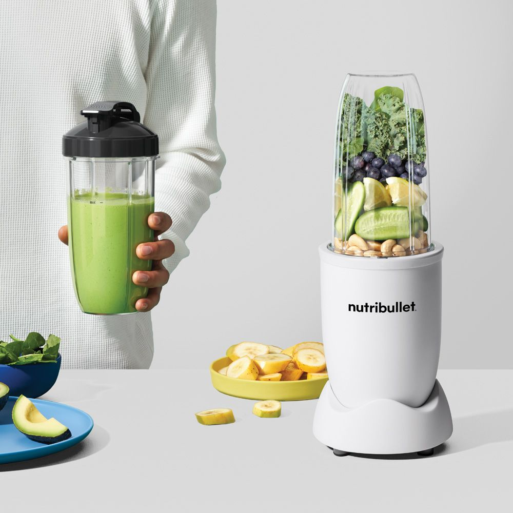 Witte Nutribullet met fruit. Hand houdt smoothiebeker vast. Achtergrond: avocado, banaan.