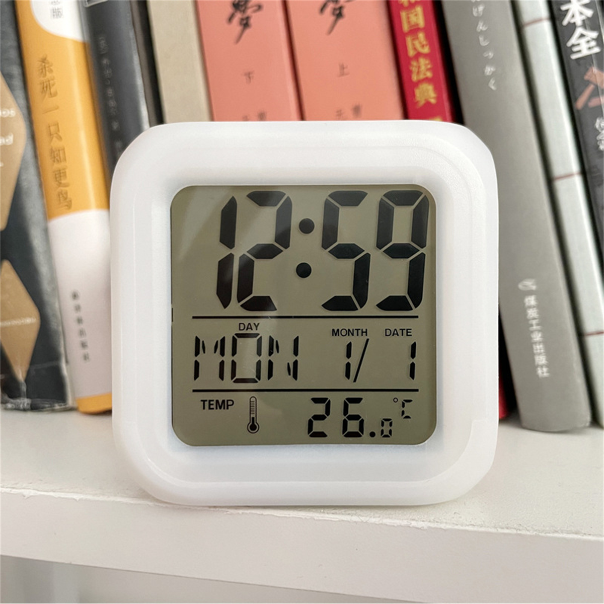 Weiße quadratische Digitaluhr mit Zeit, Datum und Temperatur. Bücher im Hintergrund.