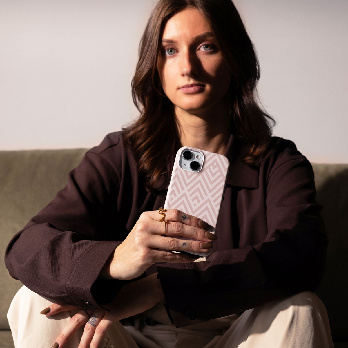 Vrouw in bruine jas en beige broek houdt een telefoon met een roze geometrische hoes vast.
