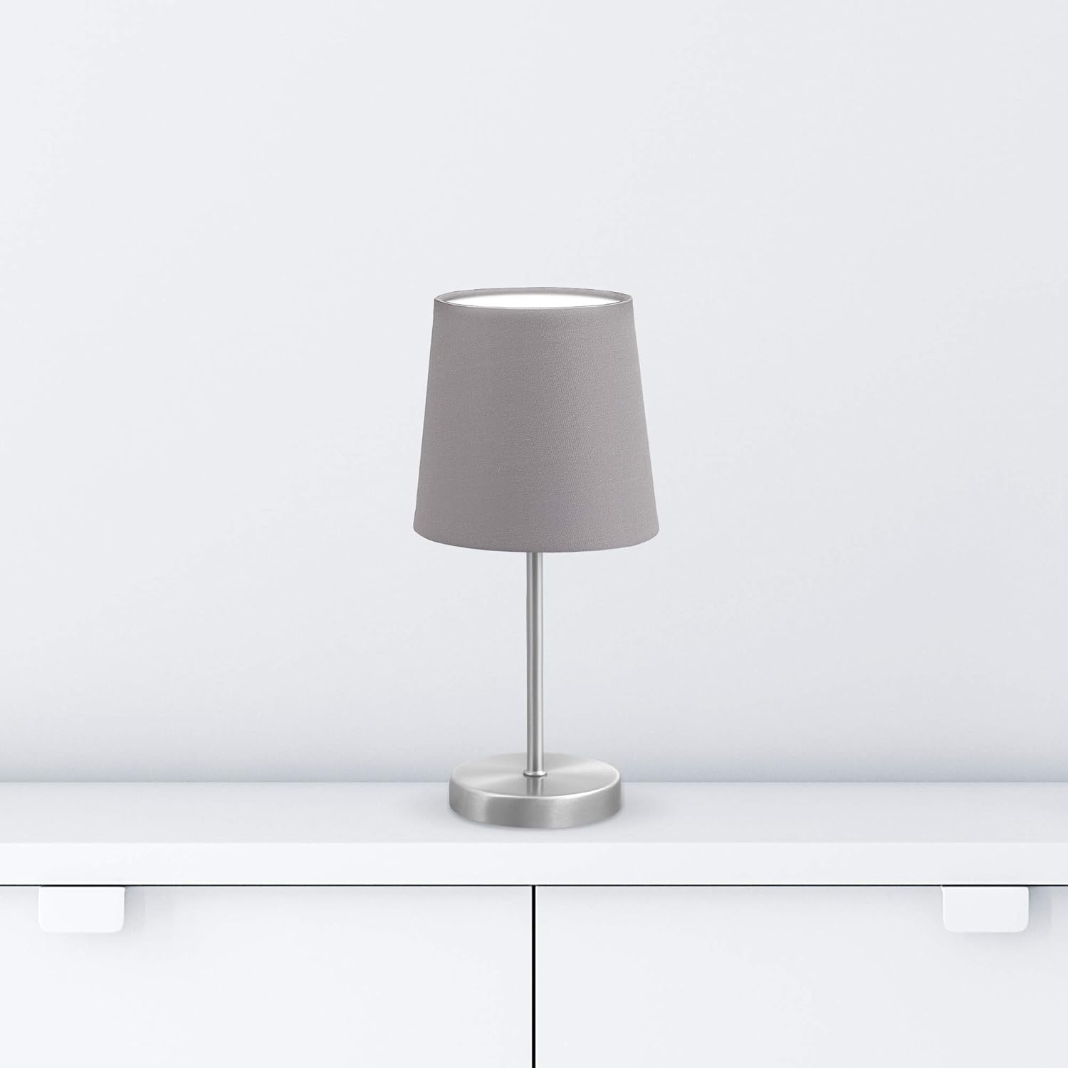 Eine graue Lampe mit Schirm steht auf einer weißen Oberfläche. Der Hintergrund ist eine weiße Wand.