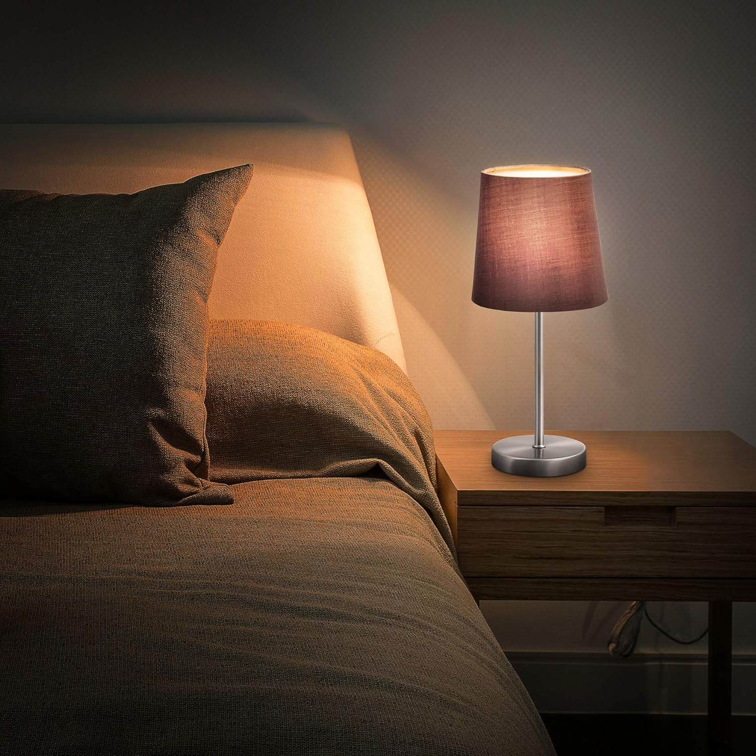 Tischlampe auf Nachttisch neben dem Bett. Lampe hat braunen Schirm und silbernen Fuß. Bett ist braun.