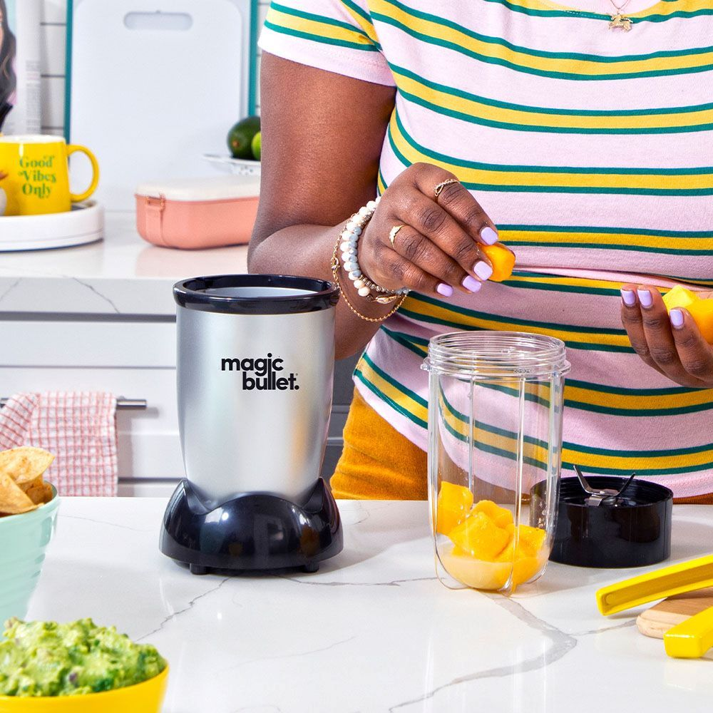 Une personne ajoutant des morceaux de mangue à un blender Magic Bullet avec d'autres ingrédients sur un comptoir.