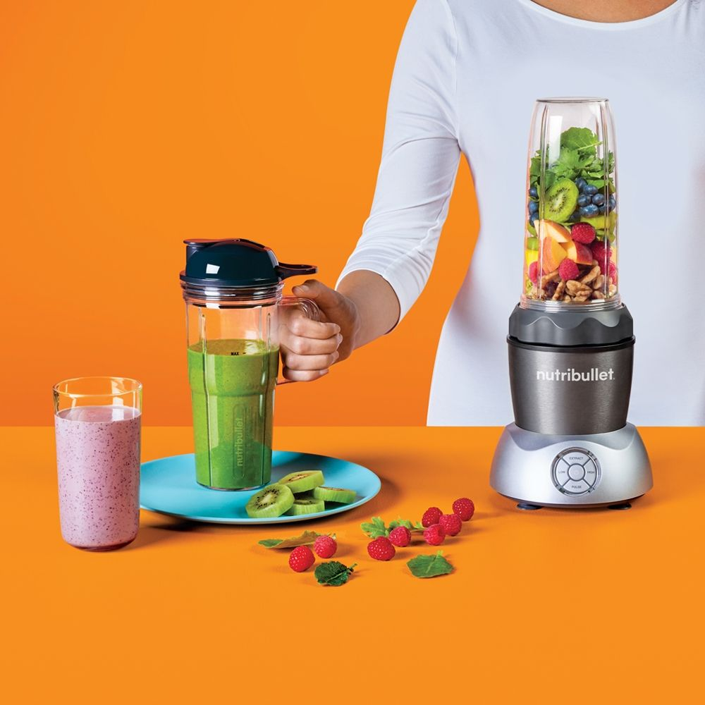 Blender met fruit, een smoothie en ingrediënten. Een persoon houdt een smoothiebeker vast.