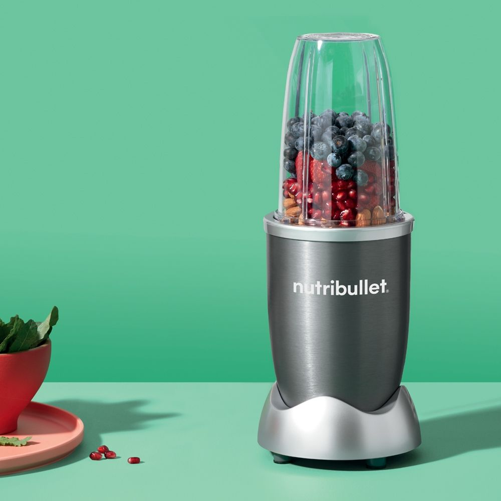Nutribullet blender met fruit erin. Groene achtergrond, rode kom en fruit.