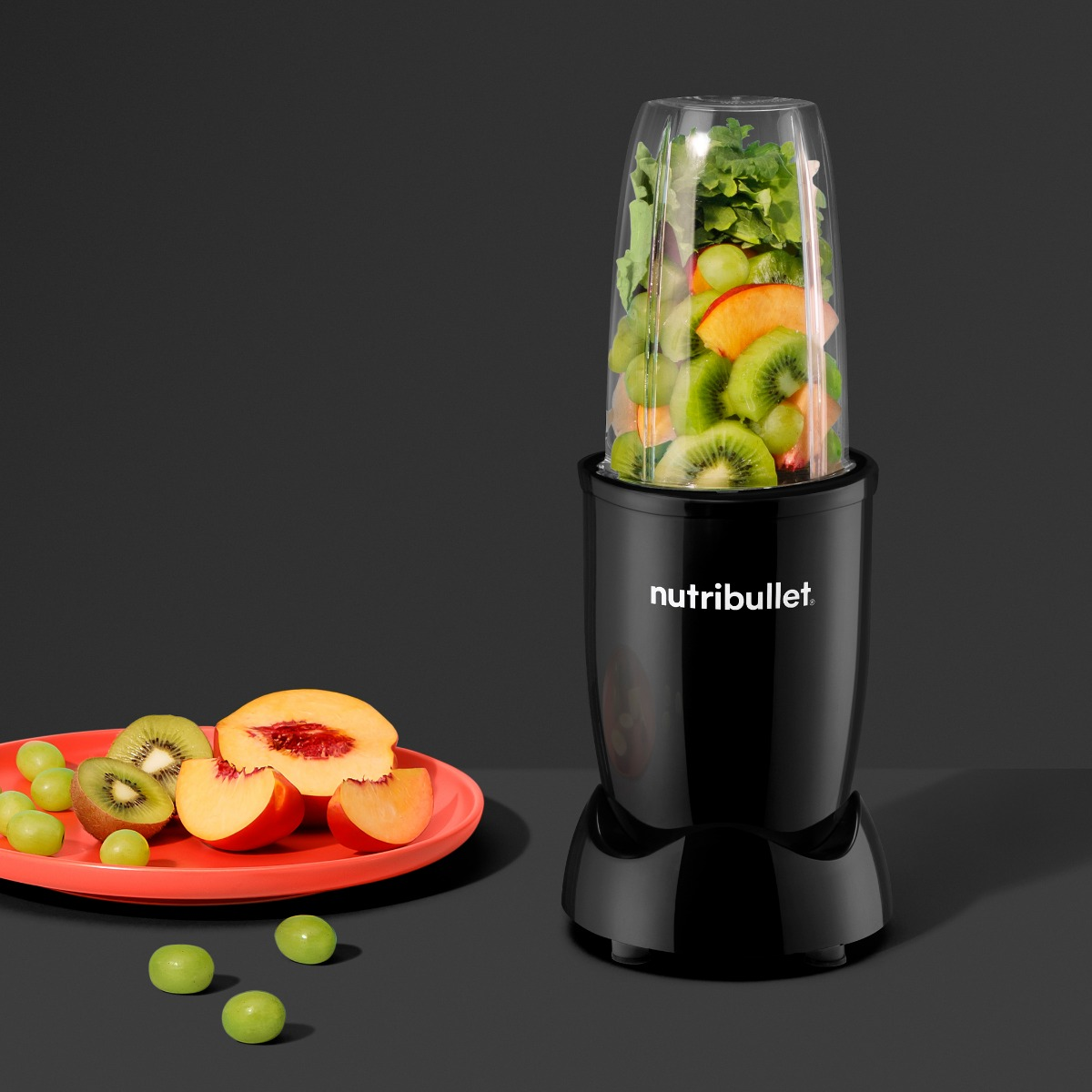 Zwarte Nutribullet-blender met fruit en groenten, rode bord, donkere achtergrond.